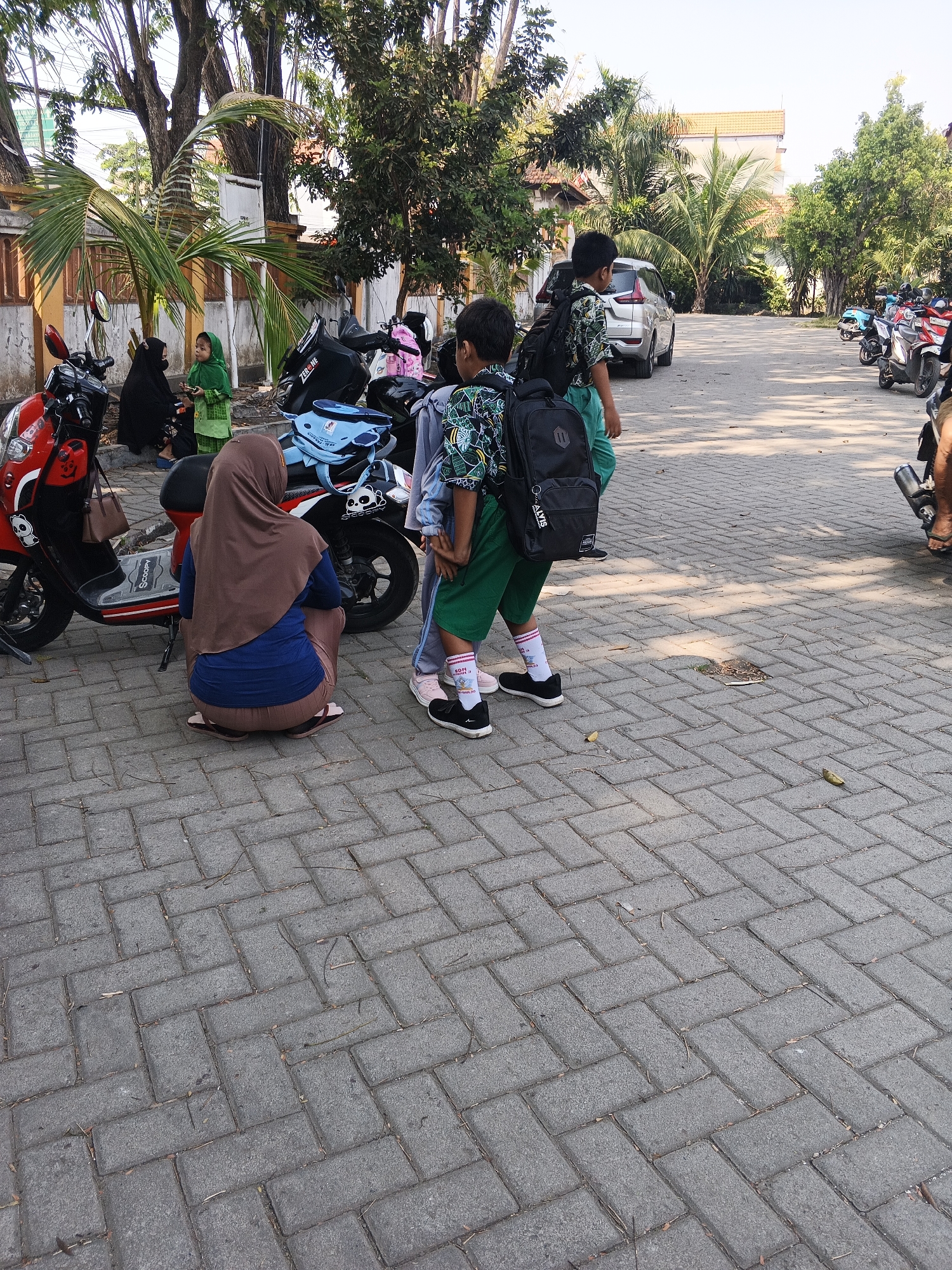 The image shows a street scene in Krian, Indonesia, with several people and motorcycles. In the foreground, a woman wearing a brown hijab and blue shirt is crouching next to a red scooter. Behind her, two young boys wearing backpacks are standing. One boy has a patterned shirt and green shorts, and the other has a lighter colored shirt. Several other motorcycles are parked in the background, as well as cars and buildings. The ground is paved with interlocking bricks. The scene appears to be taking place in a residential or school area, given the presence of children and parked vehicles.