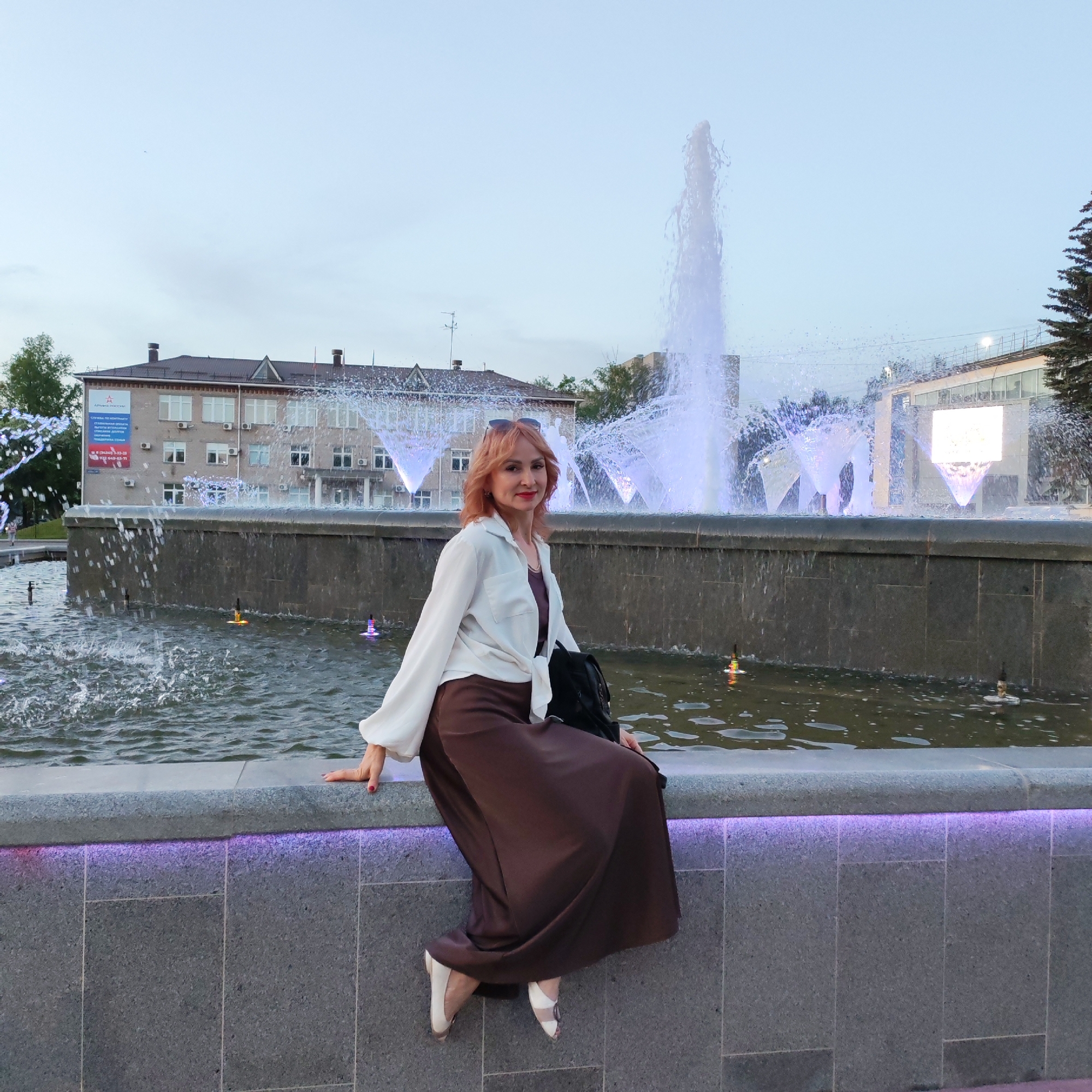 The image depicts a woman sitting on the edge of a fountain. The fountain is located in the city of Chaikovsky, Russia, in front of a multi-story building. The woman has red hair and is wearing sunglasses, a white shirt, and a brown dress. The fountain is lit with purple lights.