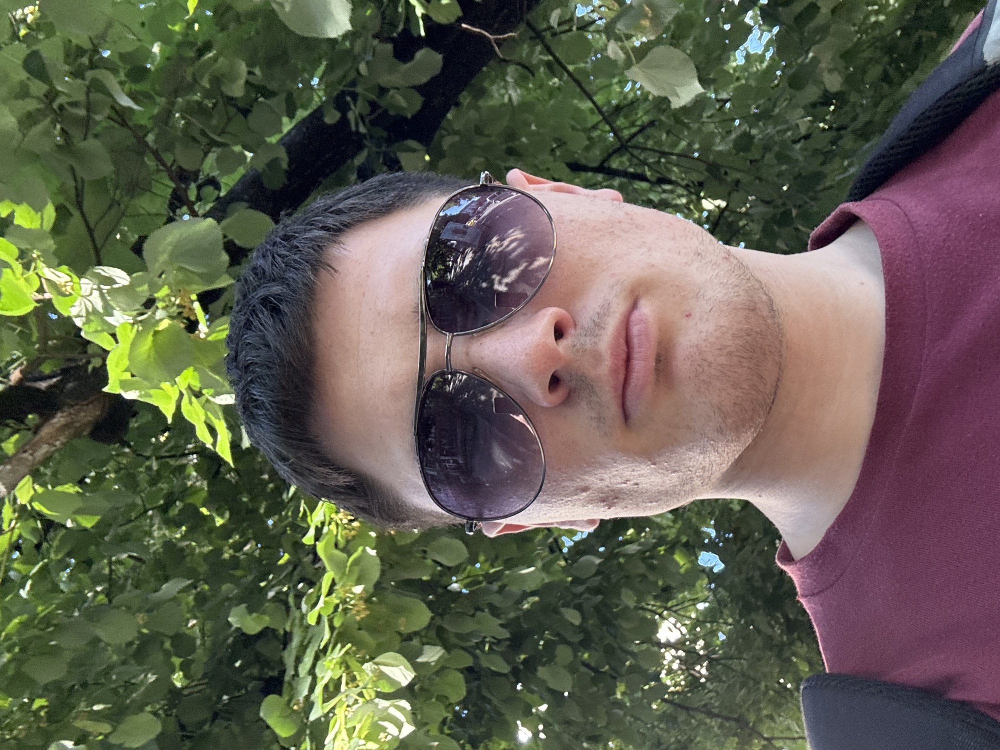 A close-up, chest-up selfie of a young man wearing aviator sunglasses and a maroon t-shirt is taken outdoors, with lush green foliage and tree branches forming the background. The man's face is partially obscured by the sunglasses, but his skin tone is light, and he has short, dark hair. The sunglasses reflect a street scene with buildings and trees. The image appears to have been taken during the day, with sunlight filtering through the leaves. The setting suggests an urban park or a tree-lined street in Stara Zagora, Bulgaria.
