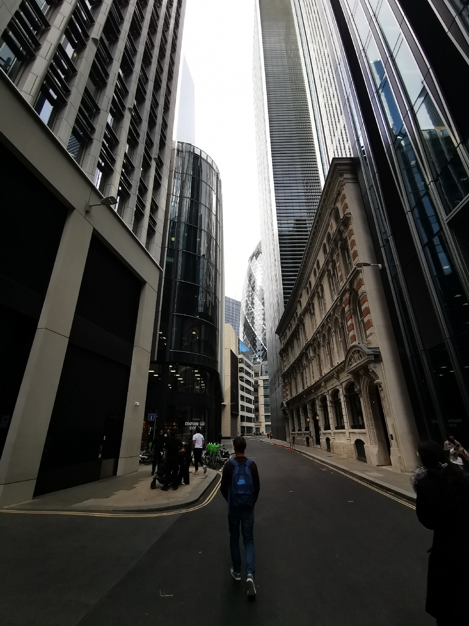 The image presents a dramatic low-angle view of a street in London, defined by a stark contrast between towering modern skyscrapers and an ornate, classical-style building. The perspective looks up the street, emphasizing the immense scale of the surrounding architecture under an overcast sky.
On the left, a modern building with a dark, textured facade and horizontal window louvers dominates the foreground. Beyond it, a sleek, curved glass building rises, followed by the distinctive diamond-patterned exterior of The Gherkin (30 St Mary Axe) in the distance, confirming the city as London.
On the right side of the street, an older, richly detailed building stands out with its beige stone facade, elaborate window arches, decorative cornices, and touches of red brick. Adjacent to it, another contemporary glass and steel skyscraper extends upwards.
The street is dark asphalt with double yellow lines. Several people are visible: in the mid-ground, a person wearing a blue Nike backpack walks away from the camera. To the left, a group of individuals gathers near a rack of shared bicycles. Visible text on a shopfront on the left reads "KENSINGTON COFFEE" and below it, "mouse tail coffee". A person is partially visible in the foreground on the right.
The daylight is diffused, suggesting a cloudy day. The scene captures the dense, dynamic urban environment, where historical and contemporary architecture coexist.