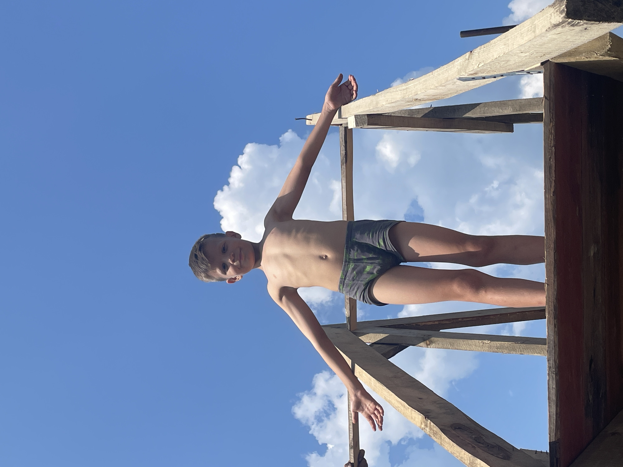 A young boy, shirtless and wearing camouflage swim trunks, is positioned on a wooden structure against a bright blue sky with scattered white clouds. He is looking towards the camera with a slight smile. His arms are outstretched, one angled upwards and the other reaching downwards along the wooden beams. The wooden structure appears to be under construction, with rough-hewn planks and some visible nails and metal fixtures. The perspective is from a low angle, looking up at the boy and the sky. The lighting suggests it is daytime, likely sunny. The overall scene conveys a sense of outdoor activity and possibly play.