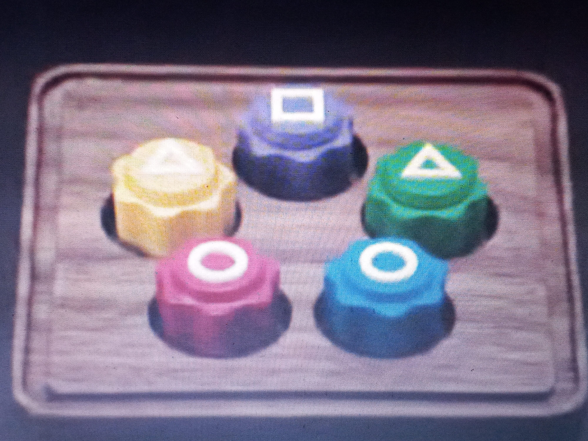 Five colorful, hexagonal cakes are arranged on a wooden tray. Each cake is topped with a geometric shape: a yellow cake with a triangle, a blue cake with a circle, a pink cake with a circle, a purple cake with a square, and a green cake with a triangle. The location is unknown.