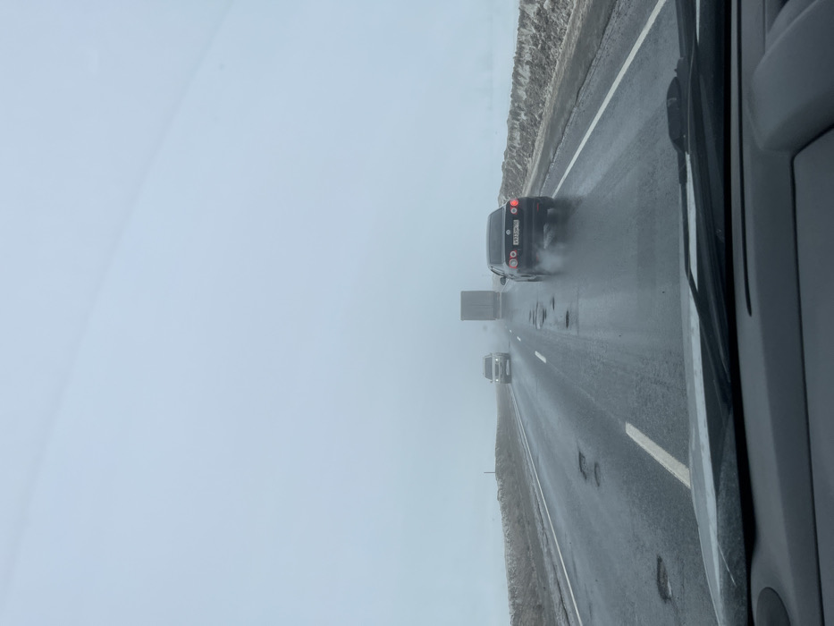 The image presents a forensic view from inside a vehicle traveling on a wet asphalt highway in or near Ivashevo, Russia, under conditions of low visibility. The sky is uniformly pale gray, indicative of heavy overcast or fog. The two-lane road features a dashed white line separating traffic directions and a solid white line on the right shoulder. Numerous significant potholes, many containing standing water, are visible in both lanes. Snow and slush banks line the right side of the road, extending into the indistinct, foggy landscape.
In the mid-ground, a dark-colored Volkswagen sedan with its brake lights illuminated (Russian license plate P933OM 90) is traveling in the right lane, leaving a trail of water spray from its tires. Ahead of the Volkswagen, in the left lane, a light-colored SUV is visible with its headlights on. Further in the distance, a large semi-truck with a light-colored trailer is partially obscured by the dense fog, also proceeding in the same direction. No people are visible. The dashboard and a portion of the windshield wipers of the observer's vehicle are present at the bottom of the frame.