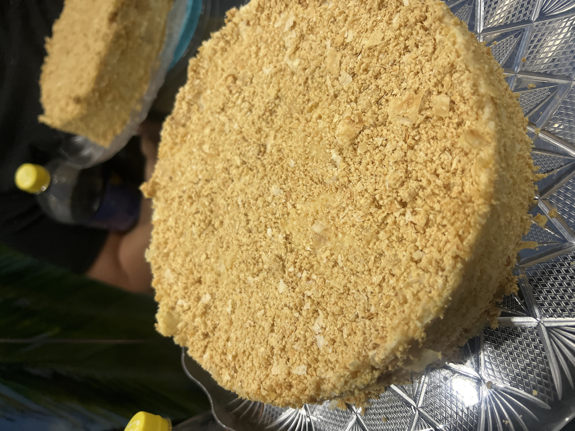 A top-down, close-up shot showcases a round cake covered in what appears to be crumbs, likely on a decorative glass plate. To the left, a portion of another cake is visible in a plastic container along with a bottle with a yellow cap. The background is dark and out of focus. There's no information to indicate that this image was taken in Budva, Montenegro.