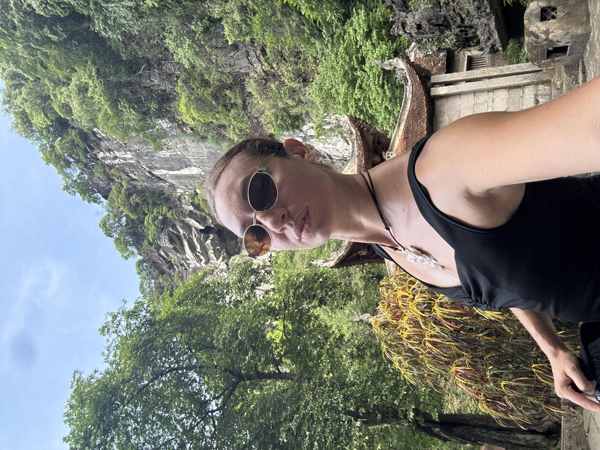 A woman is taking a selfie with a temple and a rocky, tree-covered mountainside behind her in Nam Hoa Lư Ward, Vietnam. She is wearing a black tank top and sunglasses, and a necklace with a large pendant. The woman is in the foreground, and the temple structure is visible to her right. The background consists of lush green trees and a large, imposing rock face. The sky is partially cloudy.