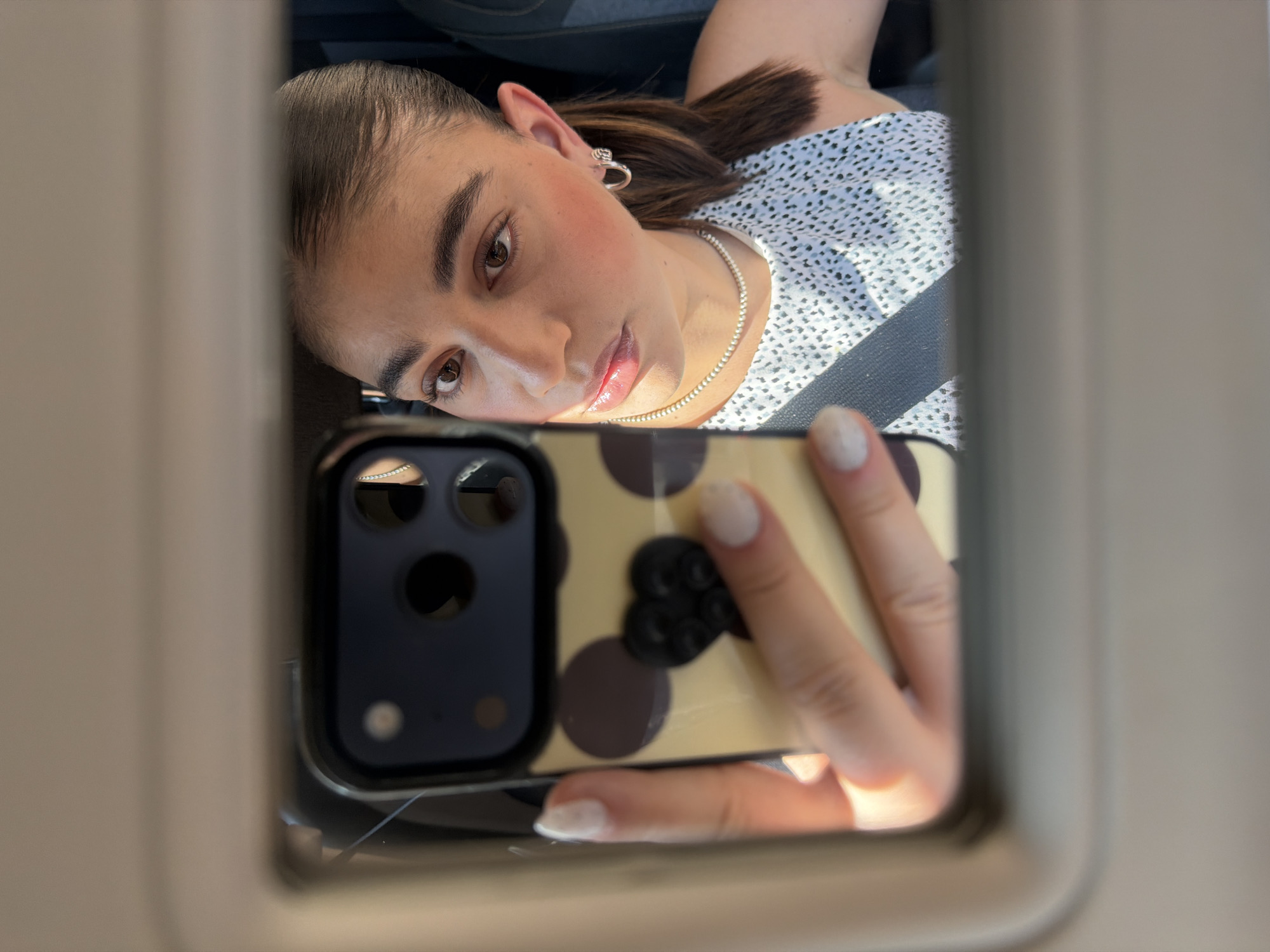 A young woman is taking a selfie in a car. She is wearing a white and black patterned shirt and a seatbelt. She is holding a phone with a black camera panel and a yellow case with brown polka dots and a black clover-shaped pop socket. She is looking at the camera with a slight smile. The image is framed by the car's sun visor. The location is not discernible from the image.
