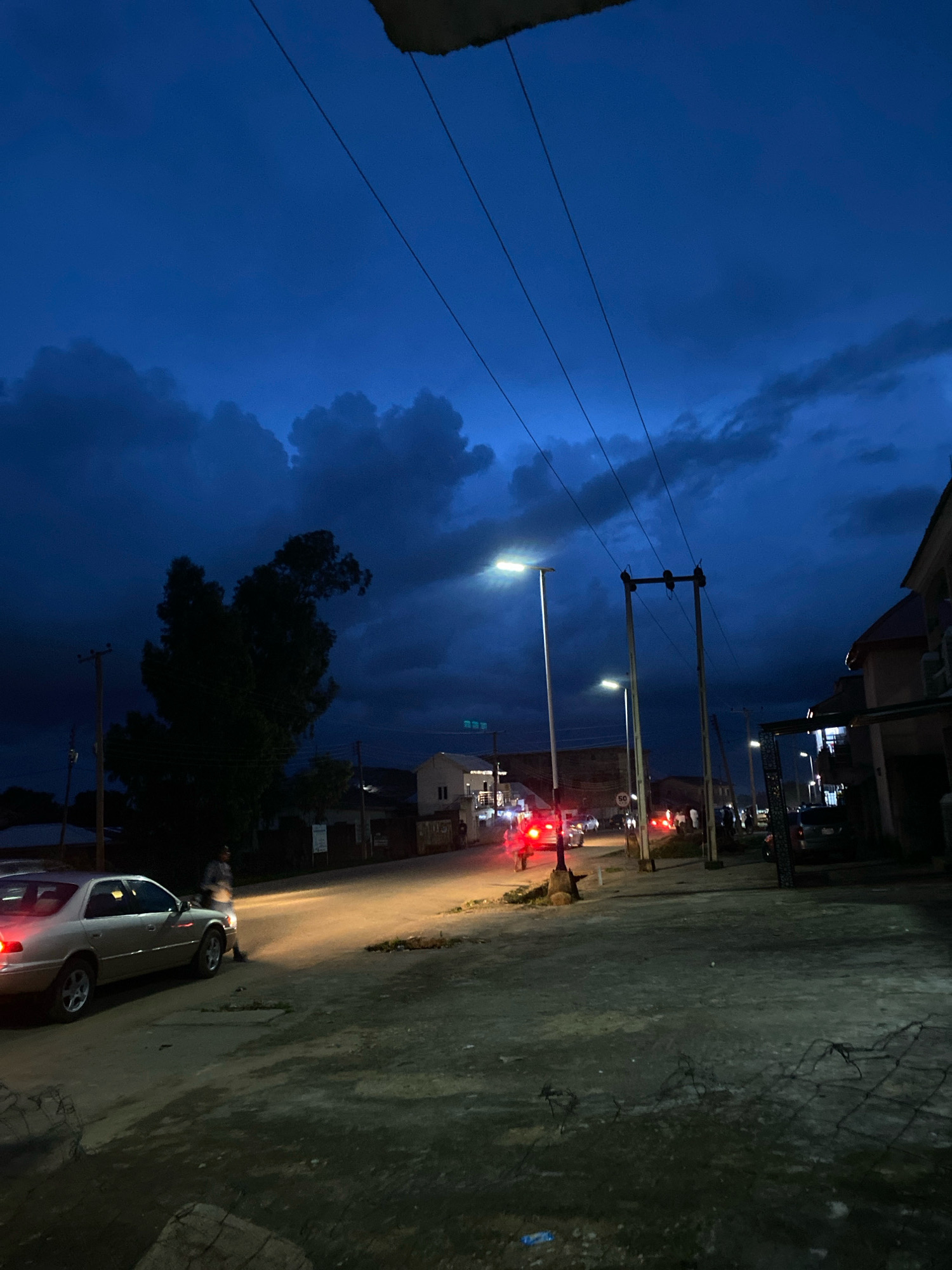 This is a low-light, eye-level shot of a street scene in Jos, Nigeria, likely captured during dusk or after dark, with a deep blue, cloudy sky overhead. In the foreground, on the left, a silver sedan is parked, its headlights illuminating the road. A person, appearing to be a man wearing a light-colored shirt and dark pants, stands near the driver's side door.
The street is paved but appears somewhat uneven, with patches of dark, wet-looking ground and some debris. Several streetlights are visible, casting pools of yellow light on the road. Further down the street, other vehicles with their taillights illuminated are present, and a few more people are standing or walking.
On the right side of the street, buildings with visible windows and structures are present, some appearing residential or commercial. Utility poles with electrical wires stretch across the upper portion of the frame, silhouetted against the dark sky. A large, dark tree dominates the left-middle ground, its dense foliage obscuring the background behind it. The overall atmosphere is quiet and slightly moody, with the artificial lights contrasting against the fading natural light.