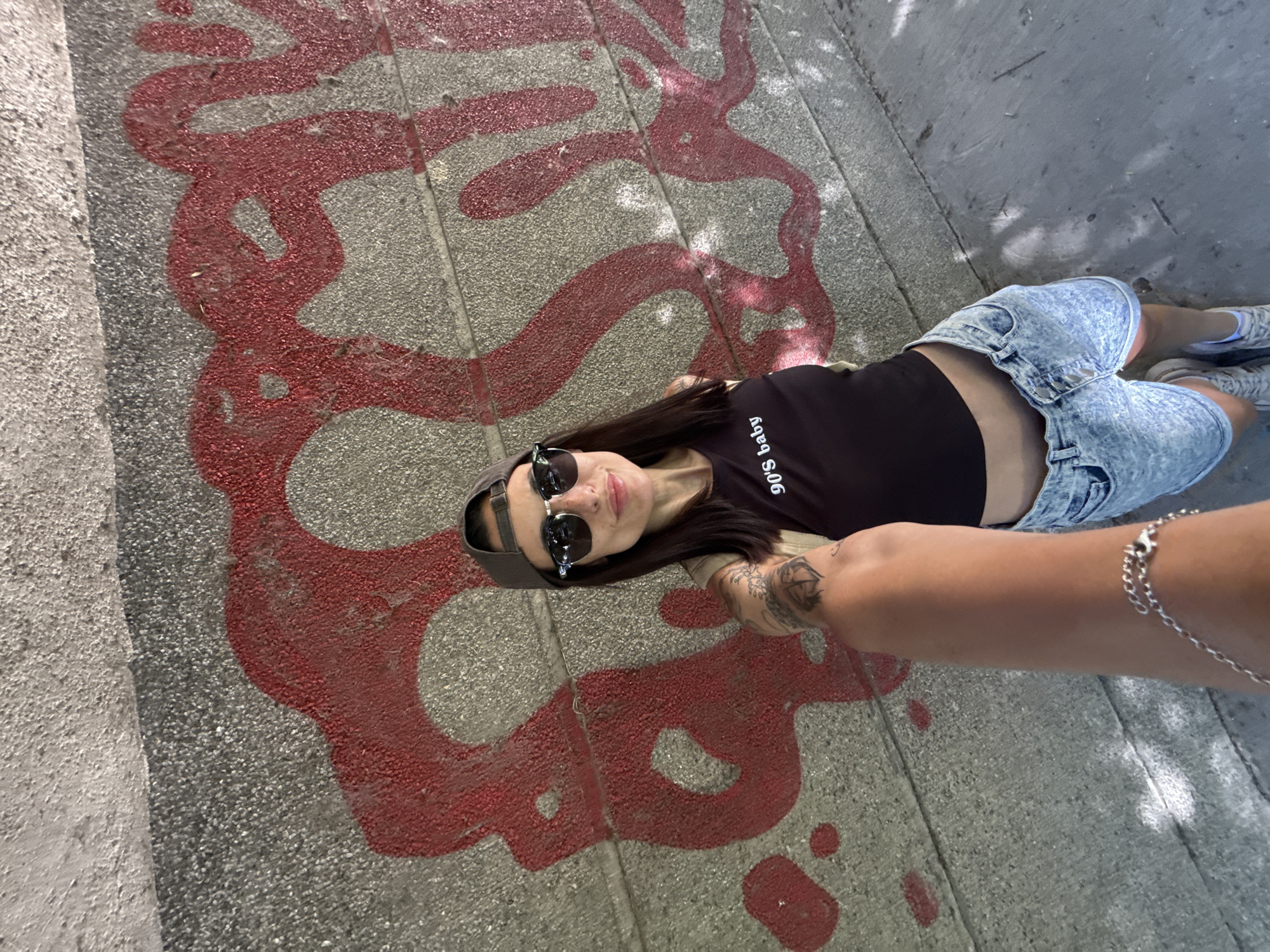 A young woman lies on a concrete surface marked with red graffiti, likely on a sidewalk or other pedestrian area. She is wearing a black top with the phrase "90's baby" printed on it, light blue denim shorts, sneakers and a backwards baseball cap. She has sunglasses on, and her arm is extended towards the camera, suggesting she is taking a selfie. A silver bracelet is on her wrist. Based on visual analysis, the location is indeterminate, and any specific reference to Pleven, Bulgaria, is not discernible from the image. The environment is urban, as indicated by the concrete and graffiti. The time of day appears to be daytime, given the lighting conditions.