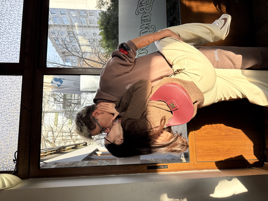 The image portrays two individuals engaged in a kiss within an interior setting, framed by a wooden window overlooking an urban landscape in Sofia, Bulgaria.
The individual on the left, with long dark hair and glasses, wears a light brown knit sweater, light beige cargo pants, and white sneakers with a dark swoosh. They are carrying a red crescent-shaped handbag with a gold-colored clasp. This individual is positioned at an angle, being lifted off the ground by the second person. Their left hand is placed on the other individual's neck/jaw.
The individual on the right, with short, spiky, light-colored hair and glasses, wears a dark brown hooded sweatshirt with a white emblem on the chest, and dark brown sweatpants featuring white "THUGBER" text on the left thigh. This individual is holding and supporting the first, with their right arm around the first individual's waist. Tattoos are visible on their right forearm.
Sunlight streams directly from the right, illuminating the subjects and casting distinct shadows on the wooden window frame and an adjacent wall. The top section of the window consists of frosted, textured glass panes. Faint mirrored text, partially legible as "d Band", is visible on the lower right window pane.
Through the clear lower windowpanes, an outdoor urban environment is visible. This includes bare deciduous trees and several multi-story, grey concrete residential buildings consistent with socialist-era architecture. A white banner featuring the distinctive blue "Billa" supermarket logo is clearly identifiable on one of the background buildings, confirming the location as Sofia, Bulgaria.
