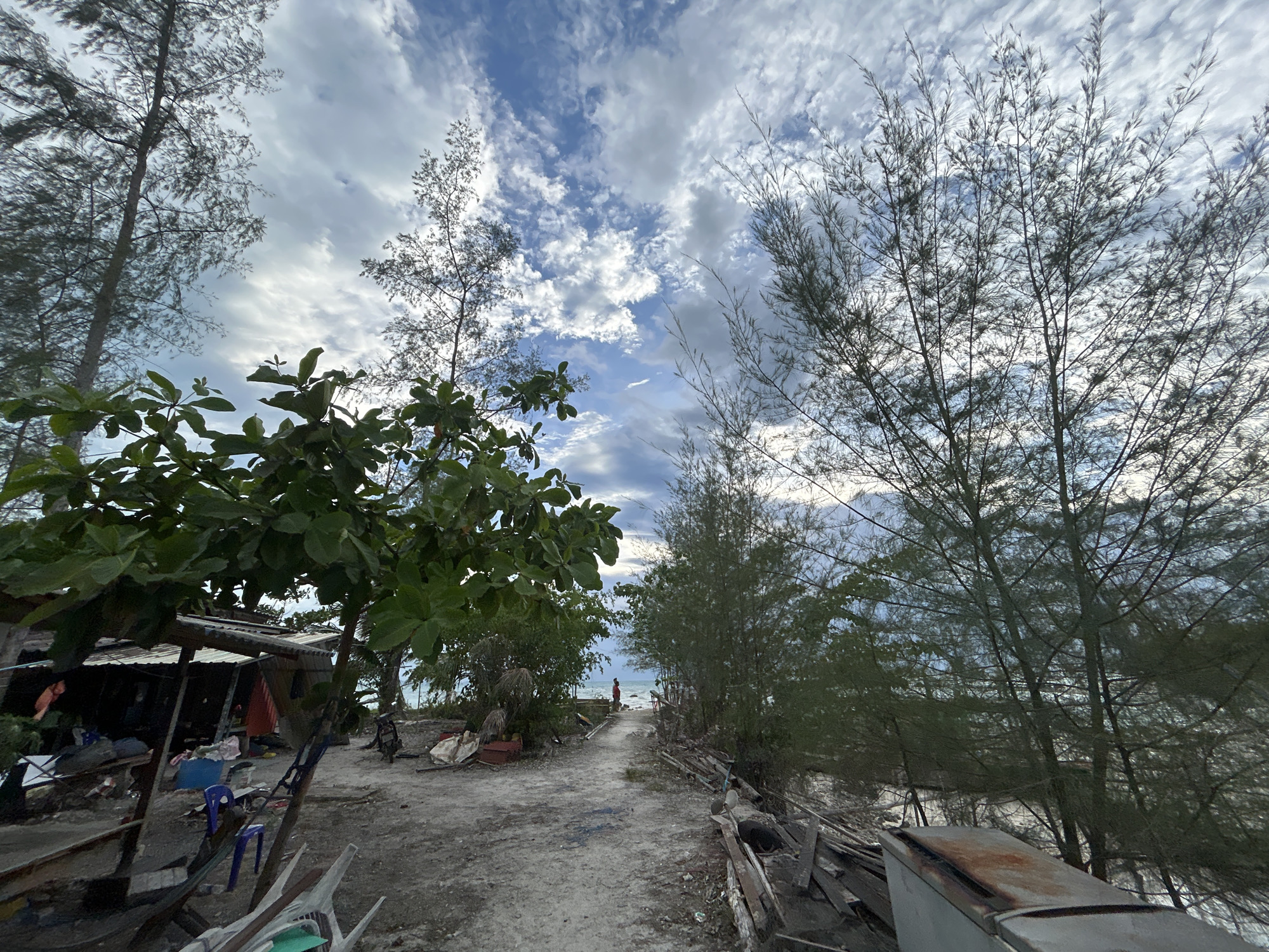 A pathway leads through sparse vegetation towards a body of water under a partially cloudy sky. On the left side of the path, a makeshift shelter constructed from corrugated metal and wood is visible, with various objects and debris scattered around it. The ground is dirt and sand. To the right of the path, there are piles of discarded lumber. In the distance, at the end of the path, a solitary figure wearing red stands facing the water. The scene appears to be a rural or undeveloped coastal area.
