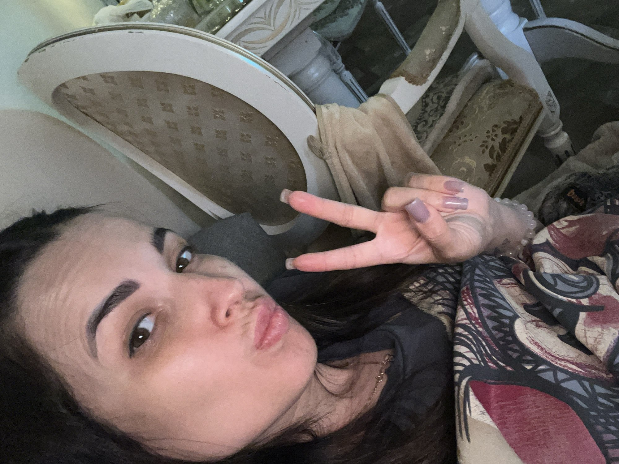 An overhead shot shows a woman taking a selfie in what appears to be a dimly lit room. The woman is in the foreground, her face is partially obscured by her hair and she is making a kissing face, with her hand raised and forming a peace sign. She has on light purple nail polish. She is wearing a dark gray t-shirt and a necklace. The background consists of a vintage-looking armchair and a table. The armchair is upholstered in a light beige fabric with a subtle pattern and has a carved white frame. The table is also white and appears to be ornate. A patterned fabric, possibly a blanket or throw, is draped over the armchair and partially covers the woman's torso.