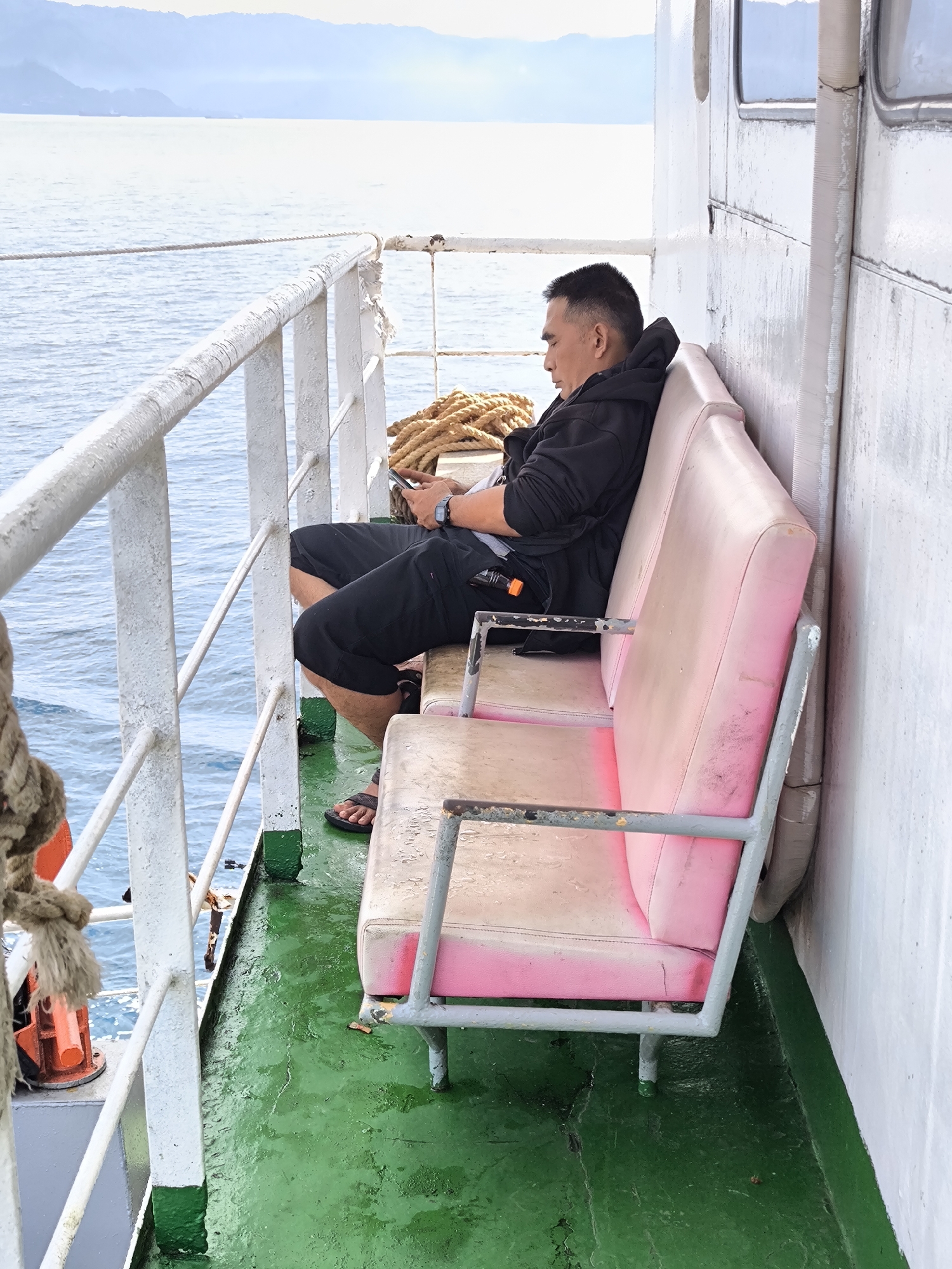 On the exterior deck of a vessel, an Asian man sits on a pink, metal-framed bench. He is in profile, facing towards the left side of the frame. The man is wearing a black hooded jacket, shorts, and sandals, and he is looking at a mobile phone in his hands. To the left of the man, a white metal railing lines the perimeter of the deck, with the sea visible beyond. A coiled rope is secured near the railing. The deck is green, and the ship's wall, to the right of the bench, is white. In the distance, land is visible under a cloudy sky. The location is Unknown, Indonesia.