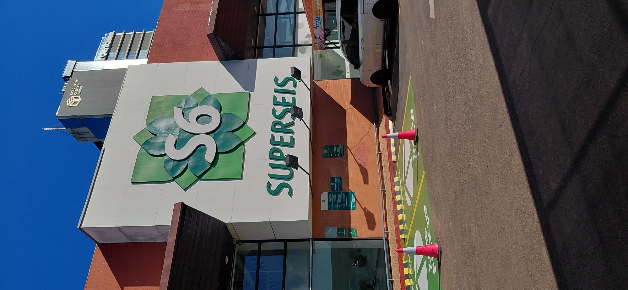 The image depicts the front exterior of a Superseis store, a prominent supermarket chain in Paraguay. The building features a white facade with a large green logo consisting of a stylized flower and the number "S6." Below the logo, the store's name, "SUPERSEIS," is prominently displayed in large green lettering. To the right of the main building, there is a modern office building with the text "EXECUTIVE CENTER OFFICE" visible. The foreground shows a parking area with designated "ZONE PICK UP" spaces marked in green and yellow. Two orange and white traffic cones are placed on the ground, and a silver minivan is parked to the right. The sky is a clear, vibrant blue. The overall scene suggests a commercial area in the city of Encarnacion, Paraguay.