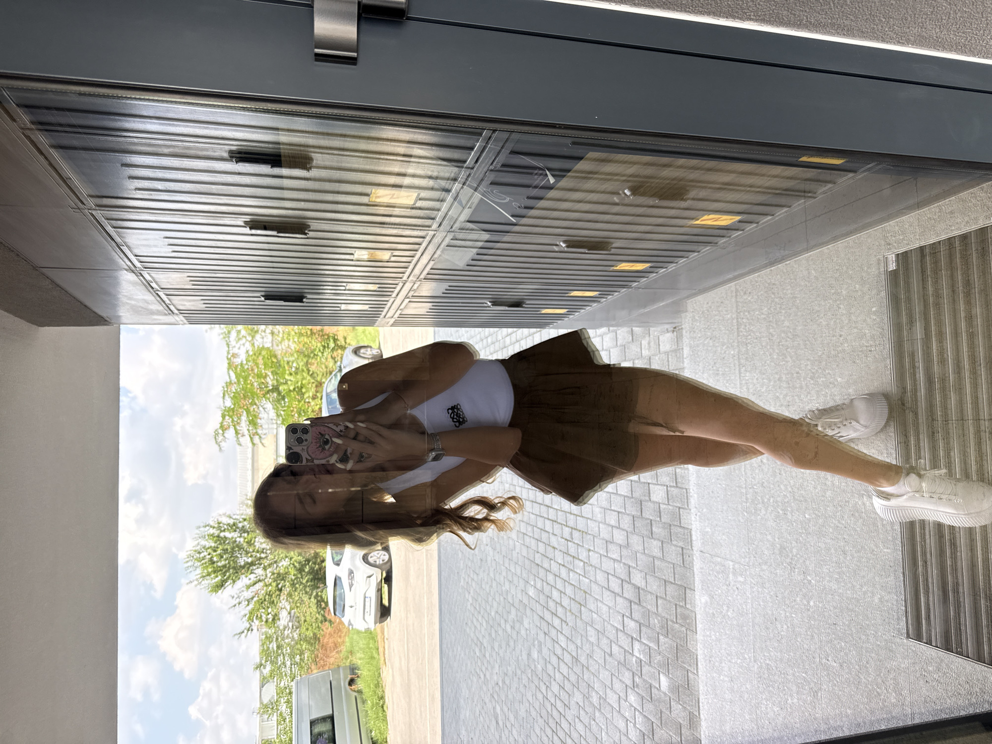 The image shows a reflection of a young woman in a glass door. She is wearing a white tank top and a brown skirt. She is holding a phone in her hand and taking a selfie. The background shows a street with cars and trees. The location is a modern building with a glass facade, which is common in urban areas. The paving stones on the ground and the style of the building suggest a contemporary urban setting. The presence of cars, trees, and a bright sky indicate an outdoor scene, likely during the daytime. The reflection in the glass creates a sense of depth and shows the surrounding environment. The overall impression is that of a casual, everyday moment captured by the woman.