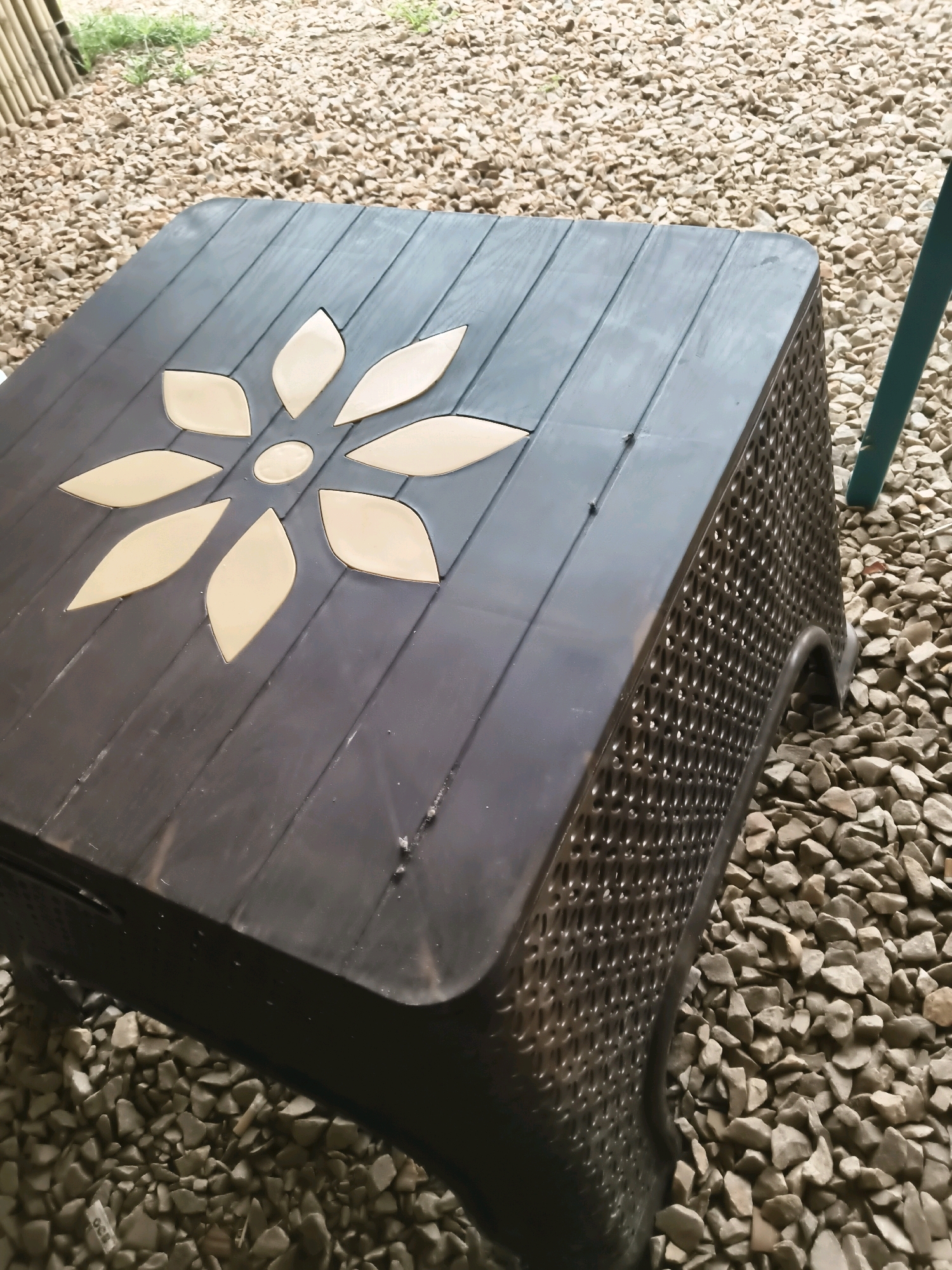 A dark brown plastic side table with a flower-shaped inlay on its surface sits on a bed of light gray gravel in Goondpur, India. The table has a textured, woven pattern on its sides and four curved legs. A teal-colored pole is visible to the right of the table. In the background, dry grass and a bamboo fence can be seen. The lighting suggests it is daytime.