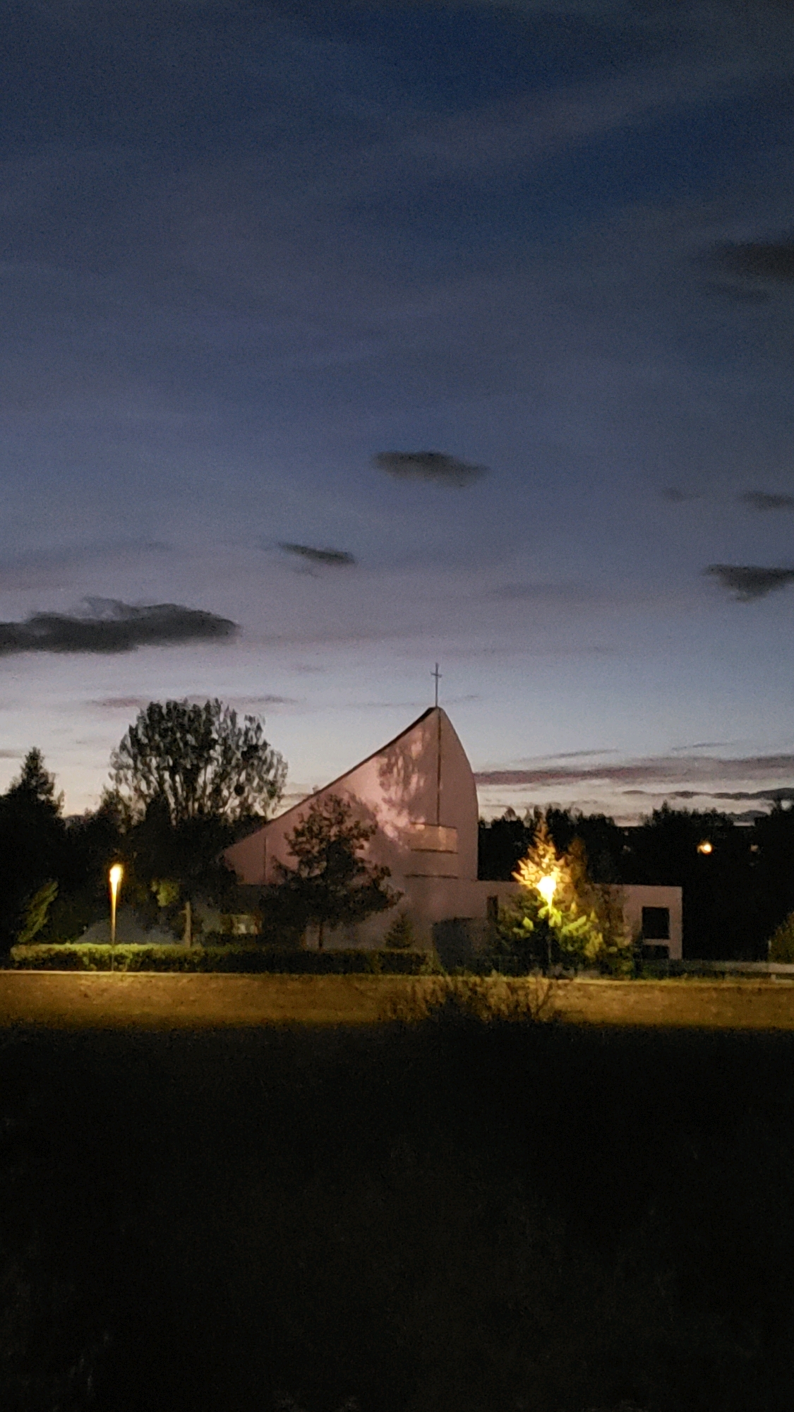 The image shows a church with a modern, angular roofline and a cross at its peak, illuminated by ambient light during twilight. The church is situated in an urban or suburban environment, with trees and other buildings visible in the background. The sky is a deep blue, with scattered clouds and hints of pink from the setting sun. A street lamp casts a yellow glow on the foreground, and some areas of the scene are shrouded in darkness.
While I cannot definitively confirm the specific location of this church within Prešov, Slovakia, based solely on the visual information, I can infer that it is a place of worship likely situated in a relatively open area to accommodate its architecture and surrounding greenery. The modern design suggests it could be a newer construction or a renovated older building. The overall atmosphere is serene and slightly mysterious due to the low light conditions.