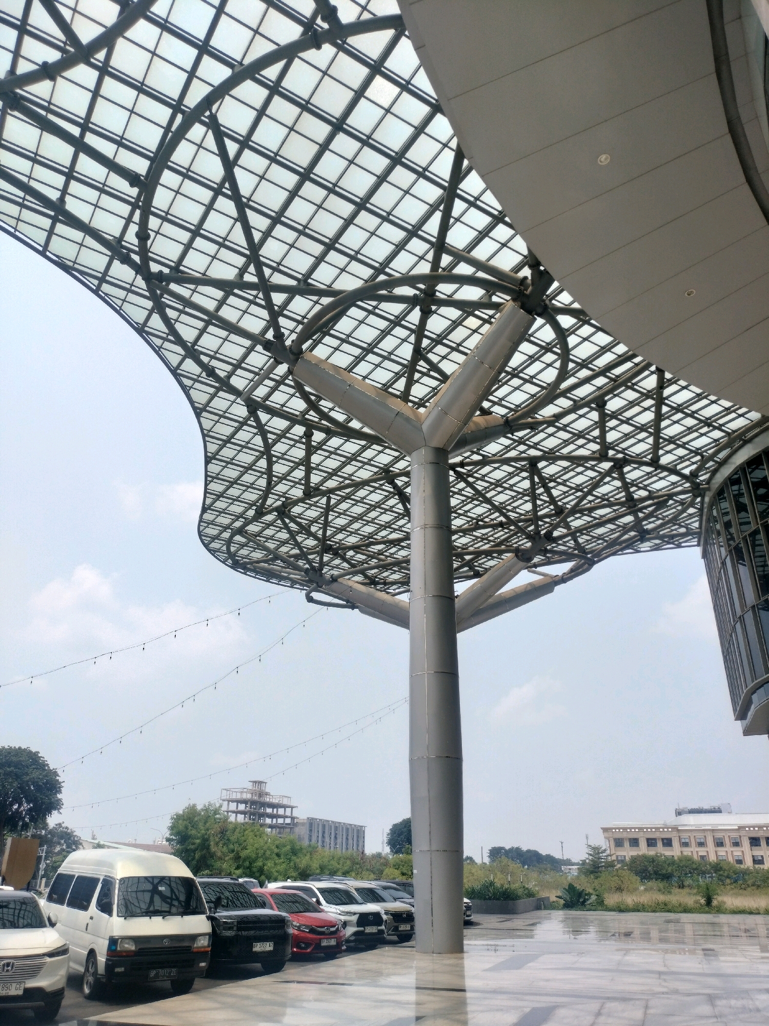 The photograph captures an outdoor scene in Batam, Indonesia, featuring a distinctive architectural element resembling a large, stylized tree supporting a modern, transparent canopy. The canopy, constructed from a grid of metal beams and translucent panels, arches gracefully overhead, providing shade. The central support structure is a large, grey, cylindrical column that branches out near the top to hold up the canopy. In the background, several cars are parked in a row, including a white van prominently featured on the left. Beyond the parking area, there are green trees and various buildings, including a multi-story structure under construction with visible scaffolding, and another building with a beige facade and numerous windows. The ground in the foreground appears to be paved with large, reflective tiles, possibly wet, indicating recent rain or cleaning. A line of small, decorative lights is strung across the sky in the mid-ground. The sky is mostly overcast with a few scattered clouds. The overall impression is of a modern commercial or public space designed with an eye for aesthetic and functional elements.