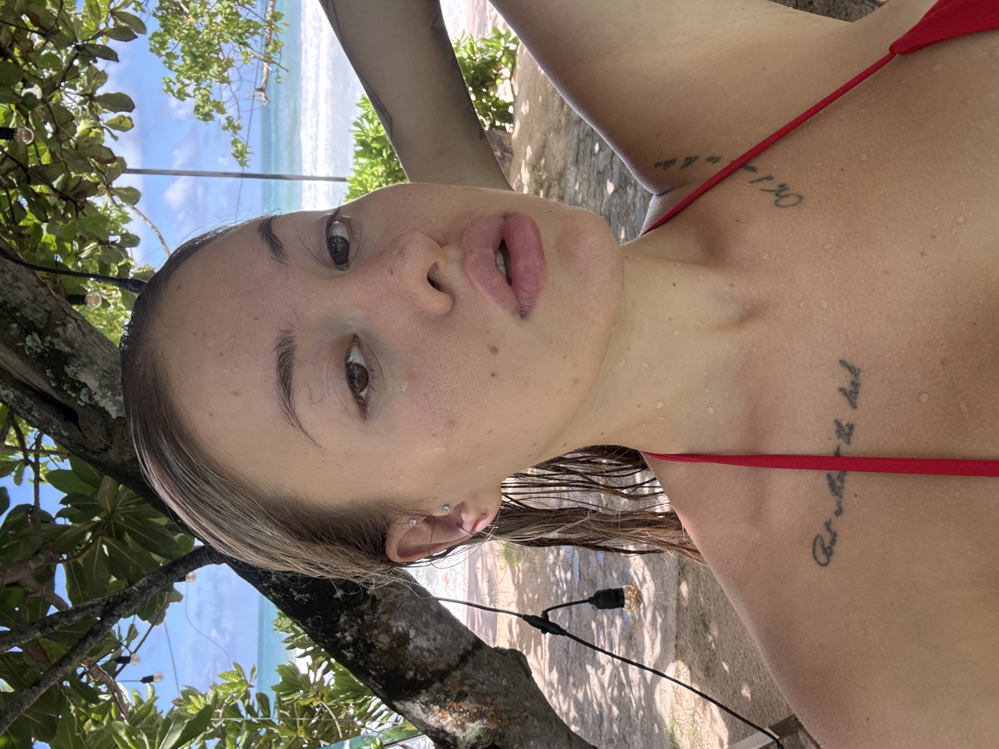 This is a self-portrait of a woman in Mirissa, Sri Lanka. She is leaning against a tree, with the ocean and beach visible in the background. The woman has wet hair and her skin is glistening with water droplets, suggesting she has recently been in the ocean or is in a humid environment. She is wearing a red bikini top. The setting appears to be daytime, with bright sunlight filtering through the leaves of the tree, casting dappled shadows on the sand. The ocean is a vibrant turquoise, and the waves are gently breaking on the shore. There are string lights with exposed bulbs visible, suggesting this might be an area set up for evening ambiance. The woman's expression is relaxed and natural. Visible on her skin are several tattoos: one on her upper chest with text that is partially obscured but seems to read "Or..." followed by some other characters, another on her left upper arm with faded text, and a smaller one on her right shoulder. The overall mood is one of leisure and enjoyment in a tropical beach setting.