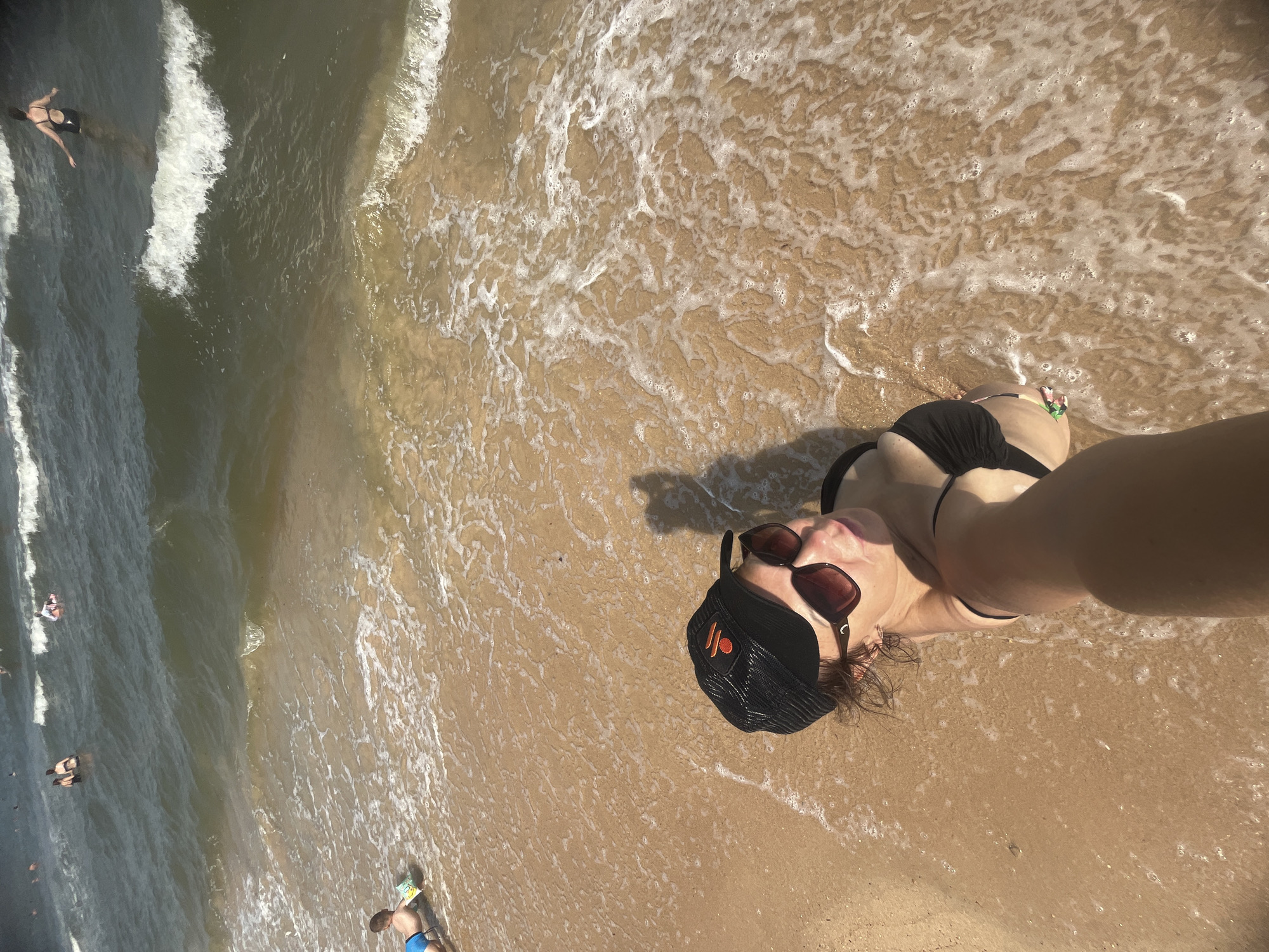 The image shows a sunny day at the beach in Golubitskaya, Russia, with people in the water and on the sand. In the foreground, a woman is taking a selfie while standing at the edge of the water. She is wearing a black bikini, sunglasses, and a black baseball cap. The water is shallow and gently lapping the shore. In the background, several people are swimming and playing in the ocean, with waves breaking on the shore. The sand is light brown and wet near the water's edge. The overall scene depicts a relaxed and enjoyable day at the beach.