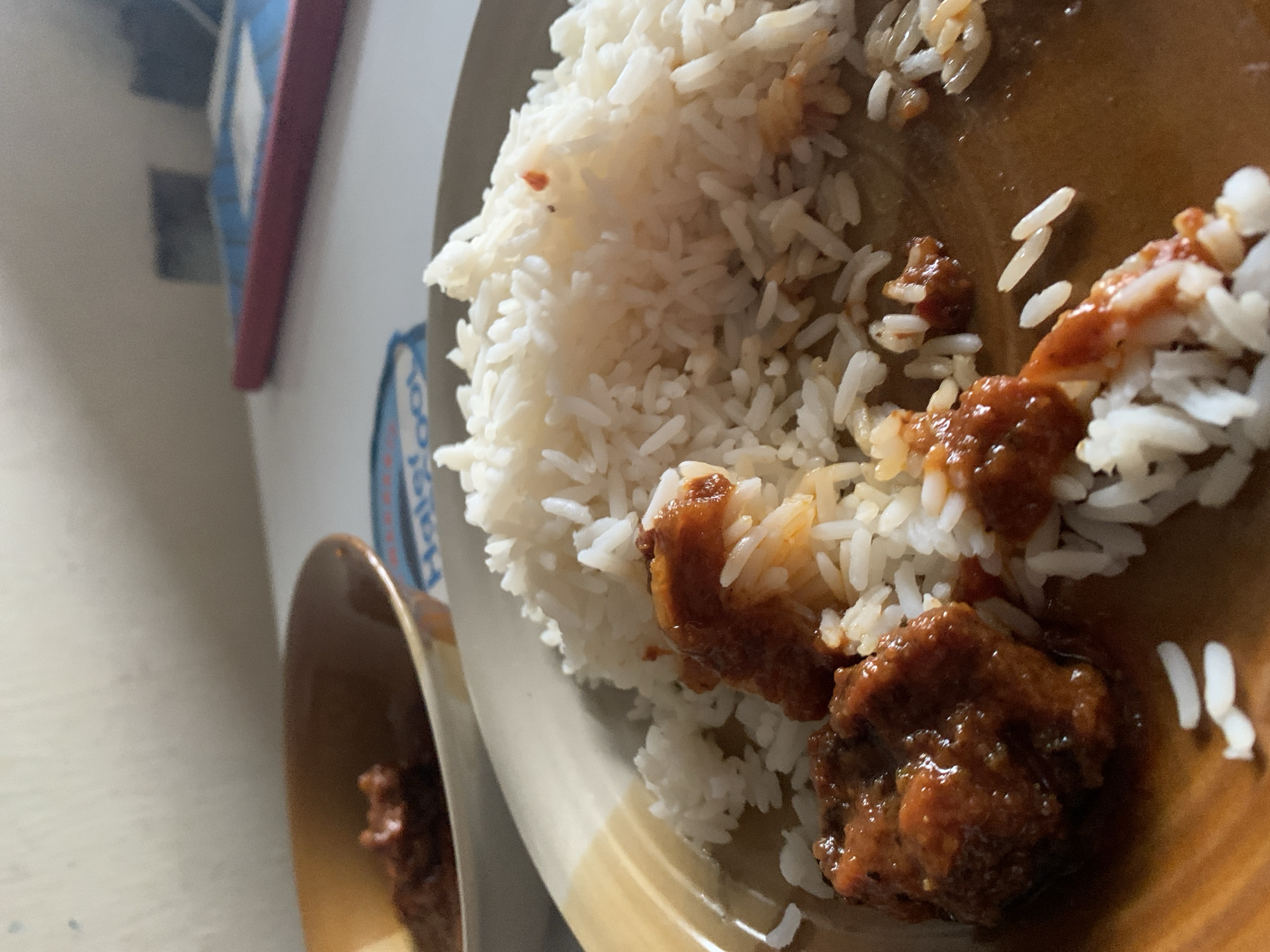 The image depicts a close-up view of a meal served on a plate, located in New Stadium, Nigeria. A light brown ceramic plate occupies the lower and central portions of the frame, holding a quantity of white, long-grain rice. Portions of a reddish-brown stew or sauce are distributed over and beside the rice, with visible pieces of a solid food item, possibly meat or protein, immersed in the sauce. The sauce exhibits a glossy texture. Some rice grains are scattered on the plate's surface, mixed with the sauce, suggesting recent consumption. In the upper left background, a smaller, similarly colored bowl contains more of the same reddish-brown stew, out of focus. The meal is placed on a light-colored table surface. To the right of the plate, a blue object with a prominent red border, possibly a tablet device or a book, rests on the table, partially out of focus. A partial logo, "Halsfoot," is visible on the table surface behind the plate. The background consists of a light, uniform wall or surface, also out of focus. No individuals are present in the scene.