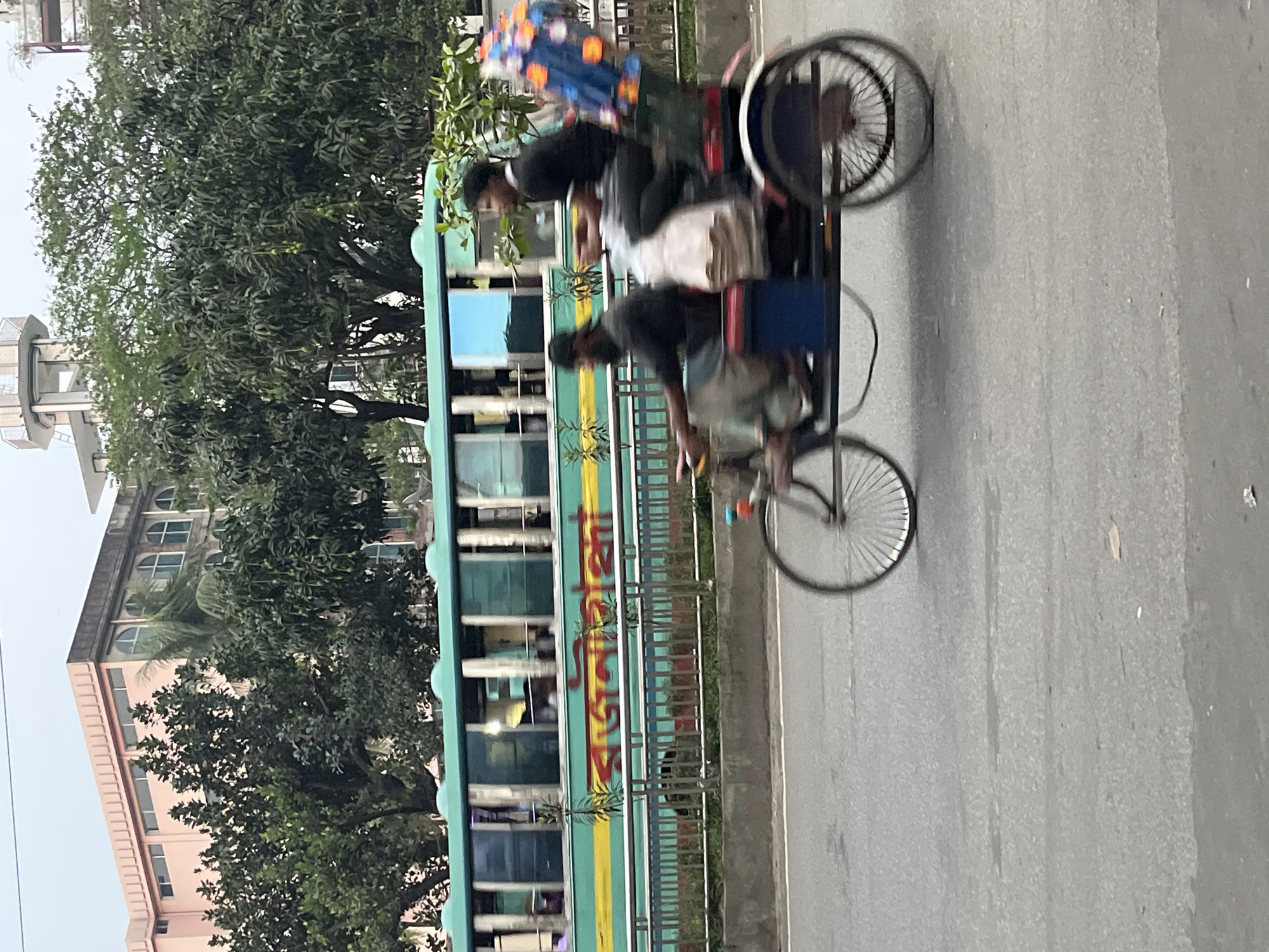 The image captures a street scene in Dhaka, Bangladesh. A cycle rickshaw, commonly used for transportation in the city, is being pedaled by a man. Another man is sitting on the passenger seat, and both figures are partially blurred, suggesting motion. Adjacent to the rickshaw, a public bus with a green and yellow exterior is moving in the same direction. Trees and buildings are visible in the background. The sky is overcast. The street appears to be paved and shows signs of wear.