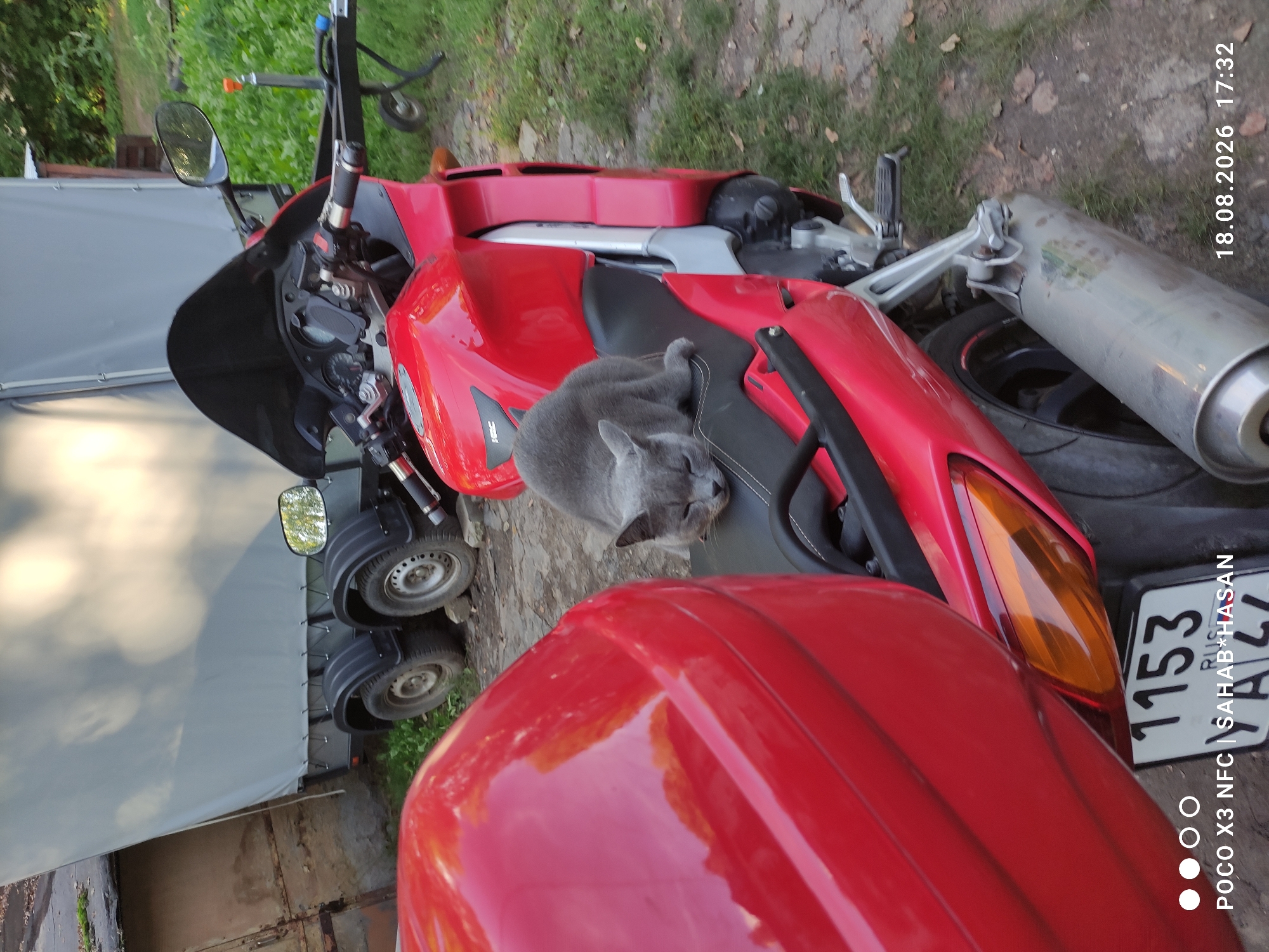 A gray cat is sleeping on the seat of a red motorcycle. The motorcycle is parked on a gravel surface next to a trailer. In the background, there is a building and some trees. The timestamp in the lower right corner indicates the date is August 18, 2026, and the time is 17:32. The text in the lower left corner reads "POCO X3 NFC | SAHAB*HASAN". The license plate on the motorcycle reads "1153 RUSSIA YA 47". The location is the city of Kostroma, Russia.