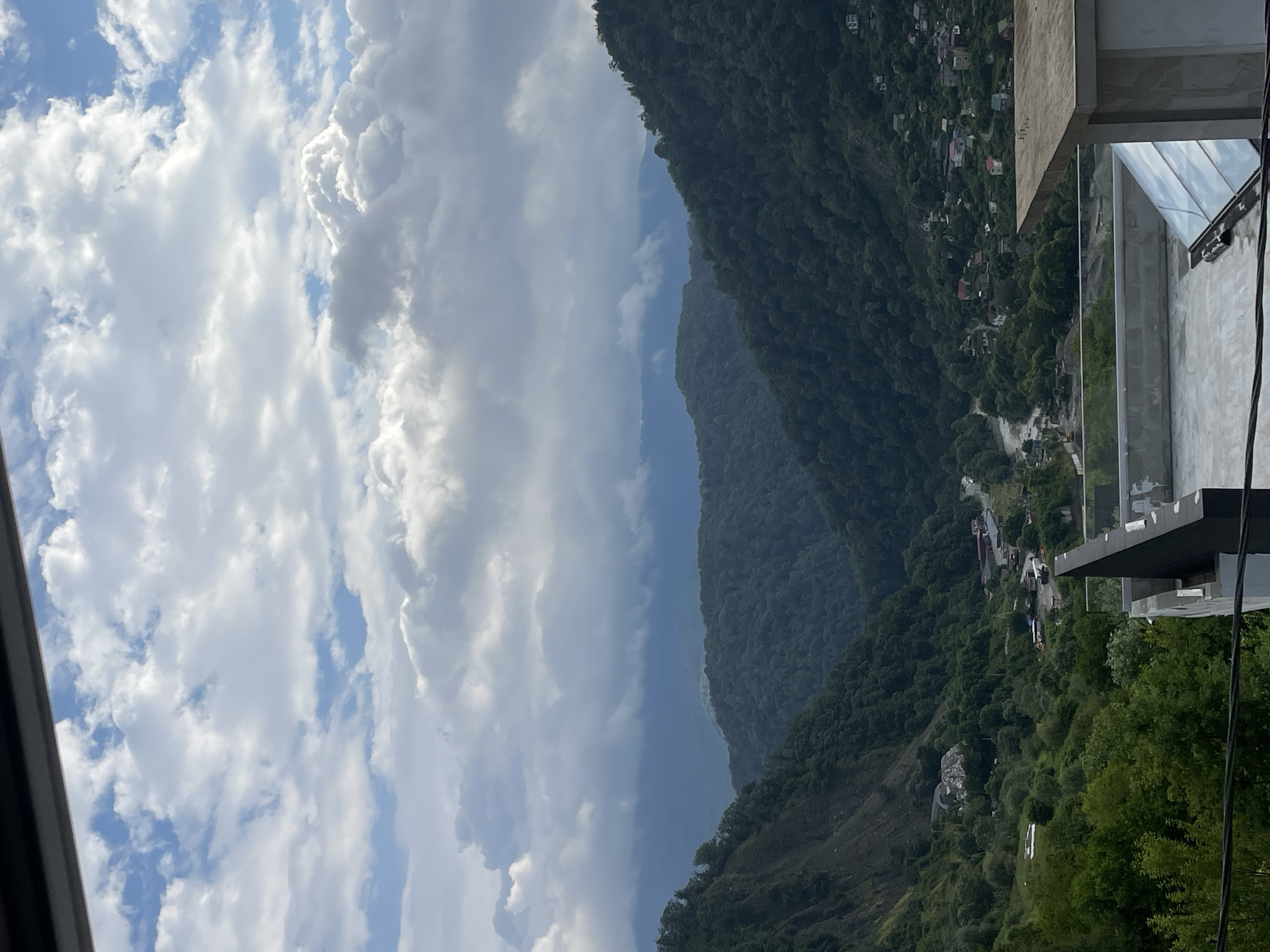 The image captures a scenic vista of a valley in Baranovka, Russia, under a dramatic sky filled with heavy, textured clouds. The foreground features a modern rooftop terrace with glass railings and a concrete structure. Beyond the terrace, a lush, densely forested valley unfolds, with rolling hills covered in dark green trees. A winding river or road is visible in the mid-ground, passing through a small settlement with numerous houses and buildings scattered across the hillsides. The slopes of the valley are steep, with some areas appearing to be rocky or undergoing some form of excavation or construction, evidenced by piles of debris and visible machinery. In the distance, more layers of mountains are shrouded in mist and the soft light filtering through the clouds, creating a sense of depth and atmospheric perspective. The overall impression is one of a tranquil, yet dynamic, natural landscape juxtaposed with human habitation and activity.