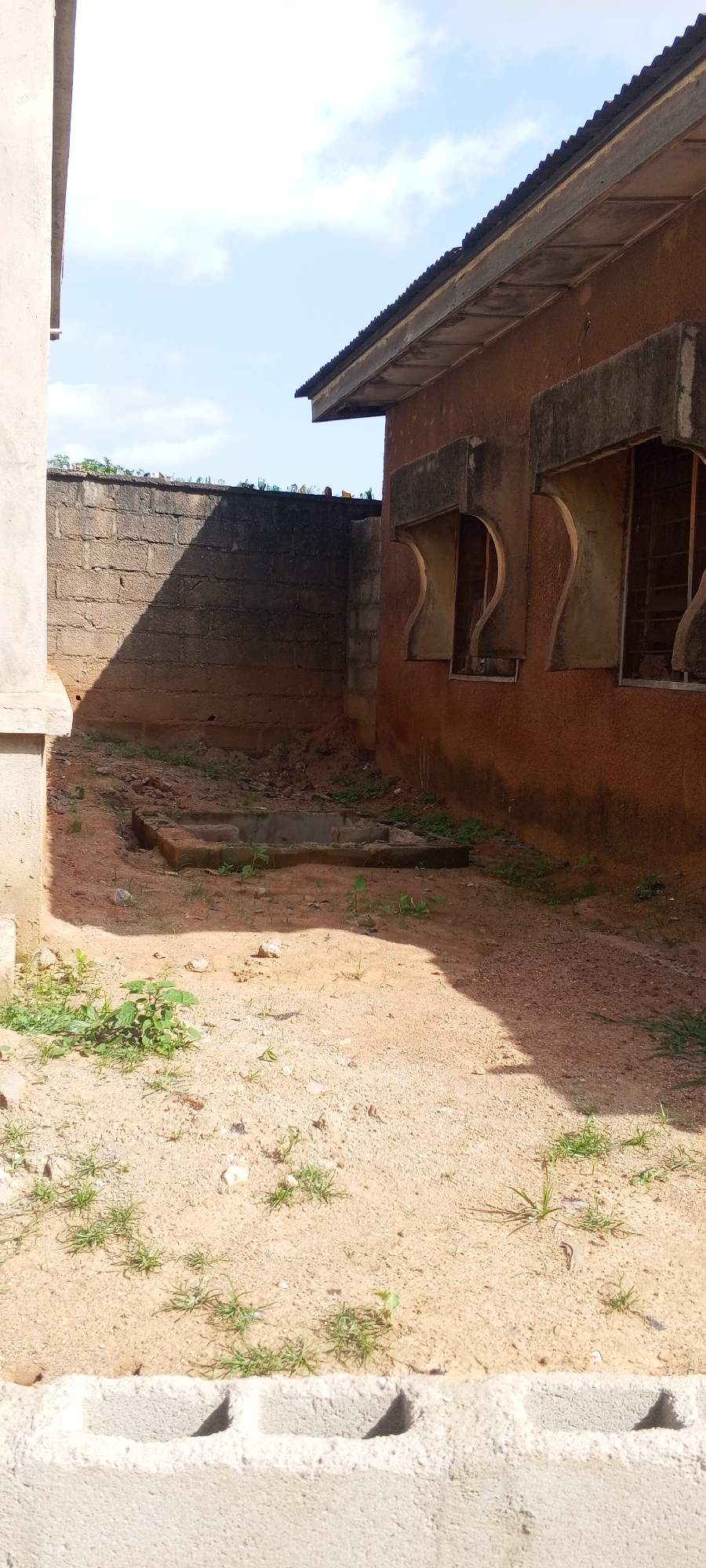 An outdoor scene in Jalingo, Nigeria, captures a dilapidated building with a corrugated metal roof and weathered orange-brown walls. Three prominent windows with unusual curved concrete frames are visible on the building's side. The ground is covered in reddish-brown sand and sparse green vegetation. A low, grey cinder block wall runs along the bottom of the frame. Behind the building, a grey brick wall extends and is topped with broken glass bottles, presumably for security. The scene is lit by natural daylight, casting shadows across the ground. The sky above is partly cloudy. The overall impression is one of neglect and disrepair.