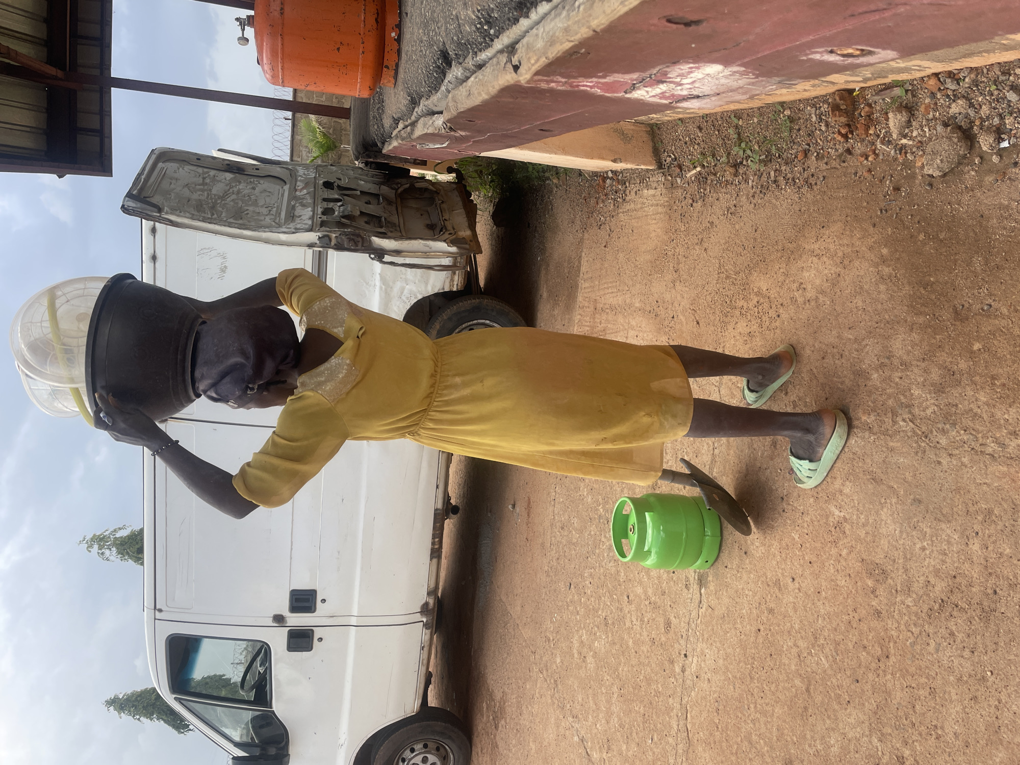 The image shows a person in a yellow dress carrying a large, clear plastic container on their head. The person is standing next to a white van with its doors open. A green propane tank and a shovel are on the ground next to the van. In the background, there are orange propane tanks, a corrugated metal roof, and a fence. The ground is made of dirt and some concrete. The sky is cloudy. The scene appears to be outdoors, possibly in a workshop or industrial area. The person's posture suggests they are carrying the container with some effort. The overall impression is one of a busy, functional setting. The crops show details of the van's interior, the surrounding ground, and the person's footwear. The time of day appears to be daytime with cloudy weather. There is no visible text in the image. The location context is Jos, Nigeria.