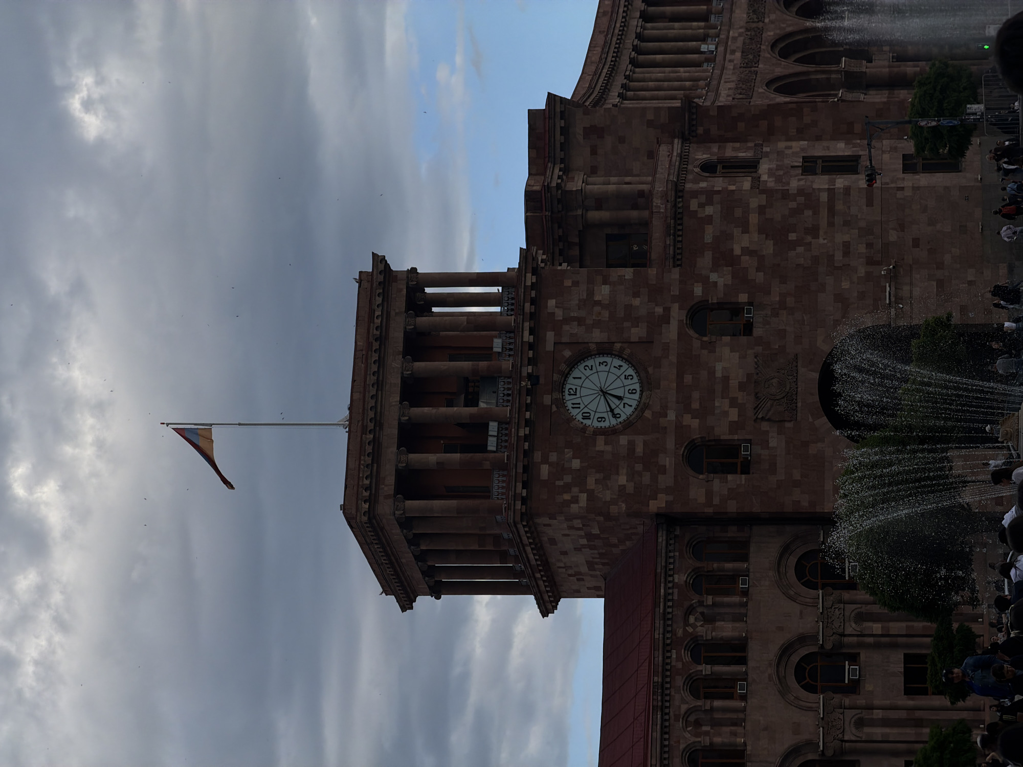 The photograph shows the Republic Square in Yerevan, Armenia, with the Government House building prominently featured. The building's architecture is characterized by its reddish-brown stonework, arched windows, and a clock tower with a large clock face. A flag of Armenia is displayed on a flagpole extending from the building. A fountain with water jets spraying upwards is visible in front of the building, with people gathered around it. The sky is partly cloudy, with varying shades of gray and blue.