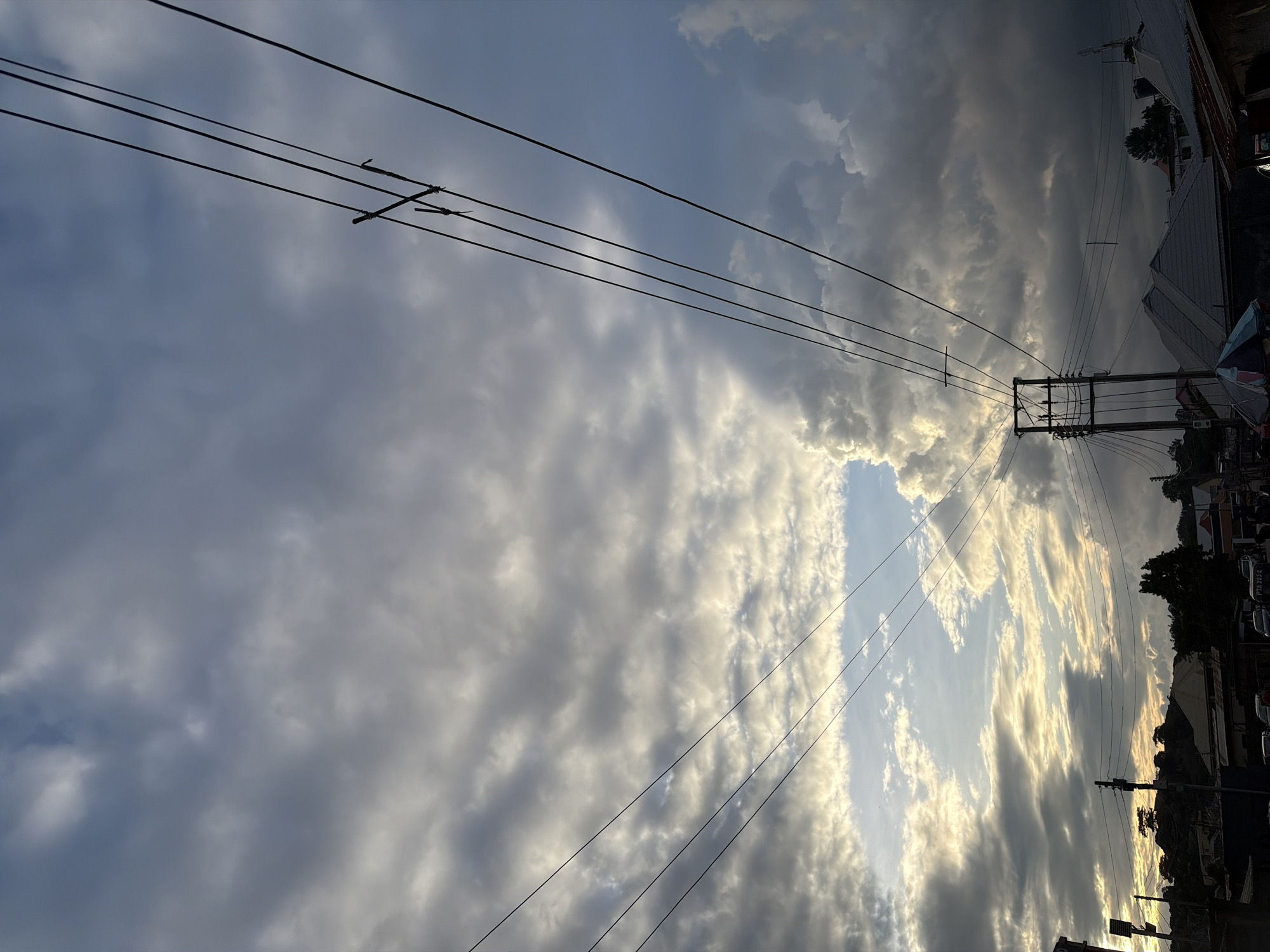 The image depicts an urban landscape in Jos, Nigeria, under a varied cloudy sky during late afternoon or early evening. The upper sky features textured altocumulus and stratocumulus clouds, appearing brighter and more diffuse on the left and central portions, revealing patches of blue sky. Towards the right, the clouds transition to a darker, more voluminous formation.
Multiple black power lines stretch diagonally across the frame from the upper left to the lower right, silhouetted against the sky. These lines are supported by at least two utility poles; one is partially visible on the upper right, and another prominent pole with cross-arms and a transformer-like structure stands in the lower central foreground.
Below the sky and power infrastructure, a settlement unfolds. On the left, a distant hill covered with sparse vegetation is silhouetted against the brighter horizon. The ground level exhibits a concentration of buildings, primarily residential, with various rooftops, including corrugated metal and flat concrete styles. Trees are interspersed among the structures. In the lower right, a street or parking area is visible, with several parked vehicles. One vehicle displays a white Nigerian license plate with the alphanumeric sequence "PTA 5252." A colorful market umbrella is partially visible in the bottom center. No distinct human figures are individually discernible.