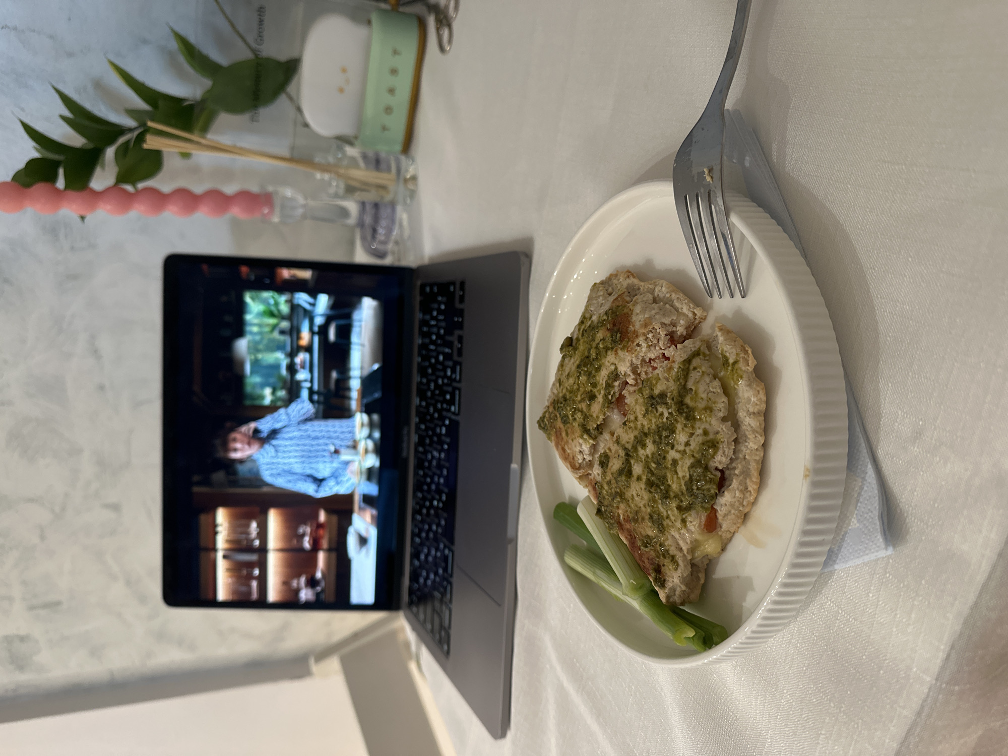 The image depicts an indoor scene focused on a meal and a laptop. A white, textured tablecloth covers the surface, upon which a white, ribbed plate holds a serving of food. The dish consists of a piece of light-colored meat or protein, possibly chicken or fish, liberally covered with a green sauce, likely pesto, and accompanied by a few segments of green onion. A silver fork rests on the rim of the plate, with a light blue napkin visible underneath.
To the left of the plate, a silver laptop is open, displaying a video on its screen. The video shows a man from the chest up, wearing a light blue patterned shirt, set against an indoor background with wooden furniture or cabinetry.
Further to the left, decorative items are present: a pink, bumpy candle and a small clear vase or bottle holding green leafy stems. Next to these, a small container has a partially visible light green label or strap that reads "TOAST." Another partial text, "Growth," is discernible on a transparent object.
The scene suggests a casual meal, possibly being enjoyed alone, with entertainment provided by the video playing on the laptop. While the provided location context is Šušanj, Montenegro, no visual cues in the image directly confirm this. The indoor lighting doesn't specify the time of day, though the candle might hint at an evening setting.