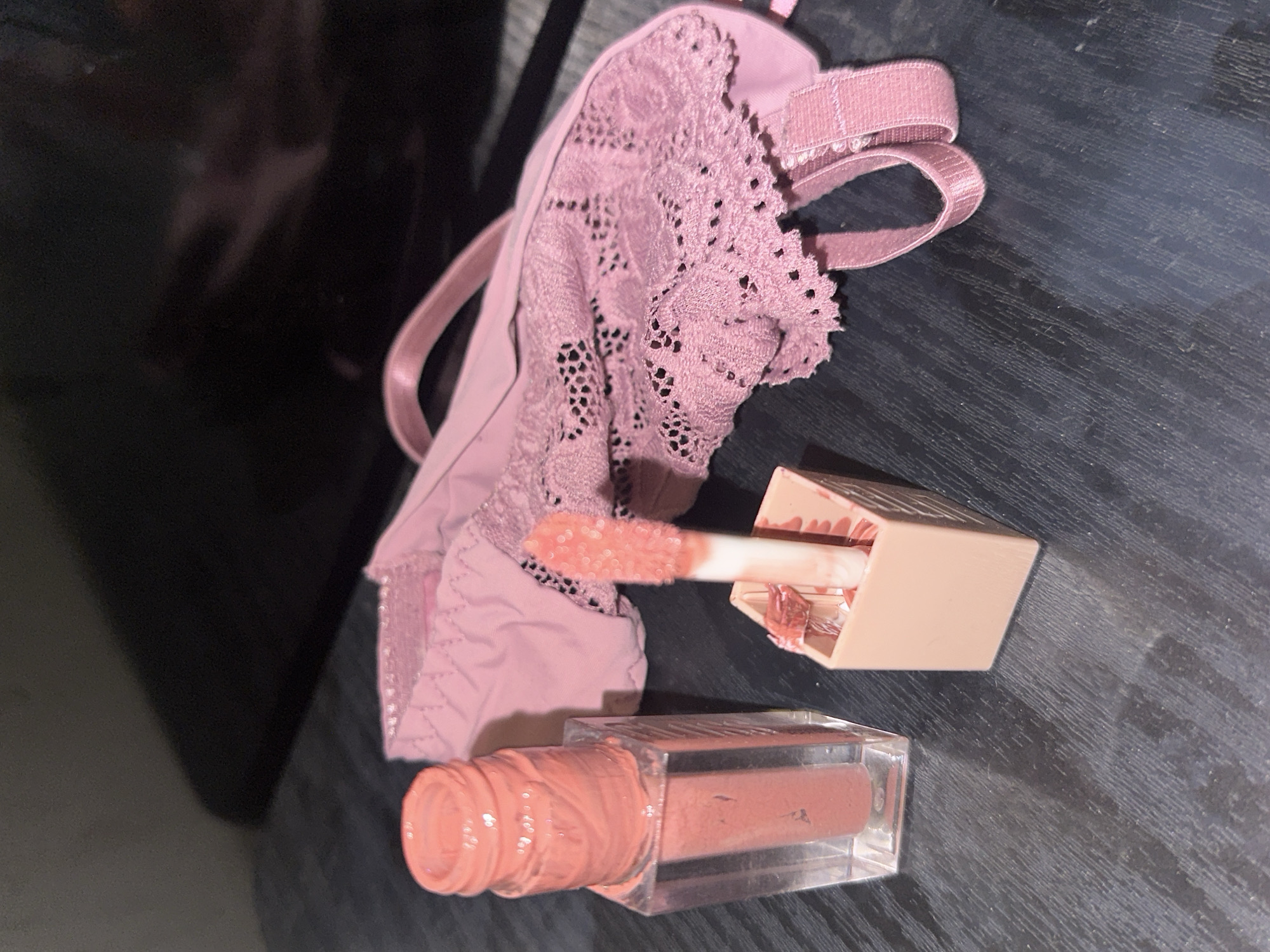 The image displays an open liquid lip product and a piece of lingerie, both resting on a dark, textured surface, likely wood, with visible linear grain patterns. The lip product, positioned on the left, consists of a clear, rectangular container filled with a coral-pink liquid. Its cap is removed, revealing the threaded neck stained with product. To its immediate right is the detached applicator wand, featuring a flocked tip coated in the same coral-pink substance, and its light peach-colored square base, which also shows internal product residue. To the right of the cosmetic items, a light mauve or dusty rose colored piece of lingerie is laid out. This garment exhibits significant lace detailing with an openwork design across its visible surface. A fabric strap, consistent with an adjustable bra or bralette strap, is visible on the right side of the lingerie. The background is dark and unfocused, indicating an indoor environment with indirect lighting. There are no discernible elements within the image that allow for the determination of a specific geographic location, including the city of Sofia, Bulgaria.
