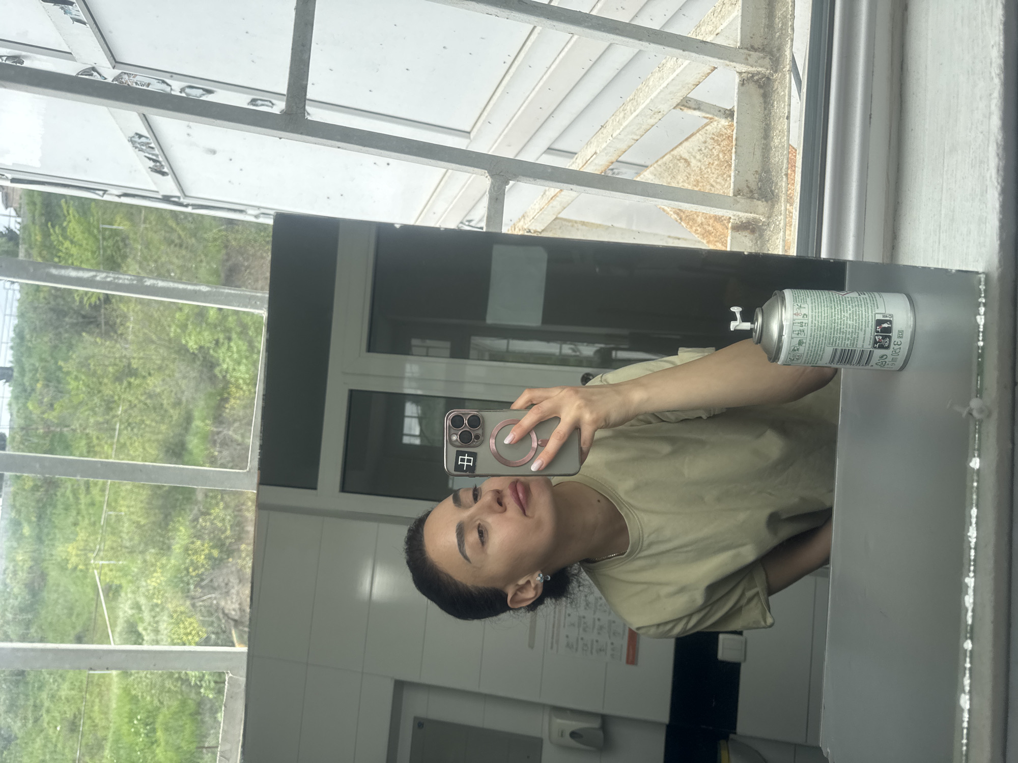 A person takes a mirror selfie in a bathroom with white tiles. A can of hairspray rests on the edge of the mirror. The reflection shows a window with a view of trees. The phone case has a Chinese character on it. Based on the architectural style and vegetation, the location could be Tbilisi, Georgia.