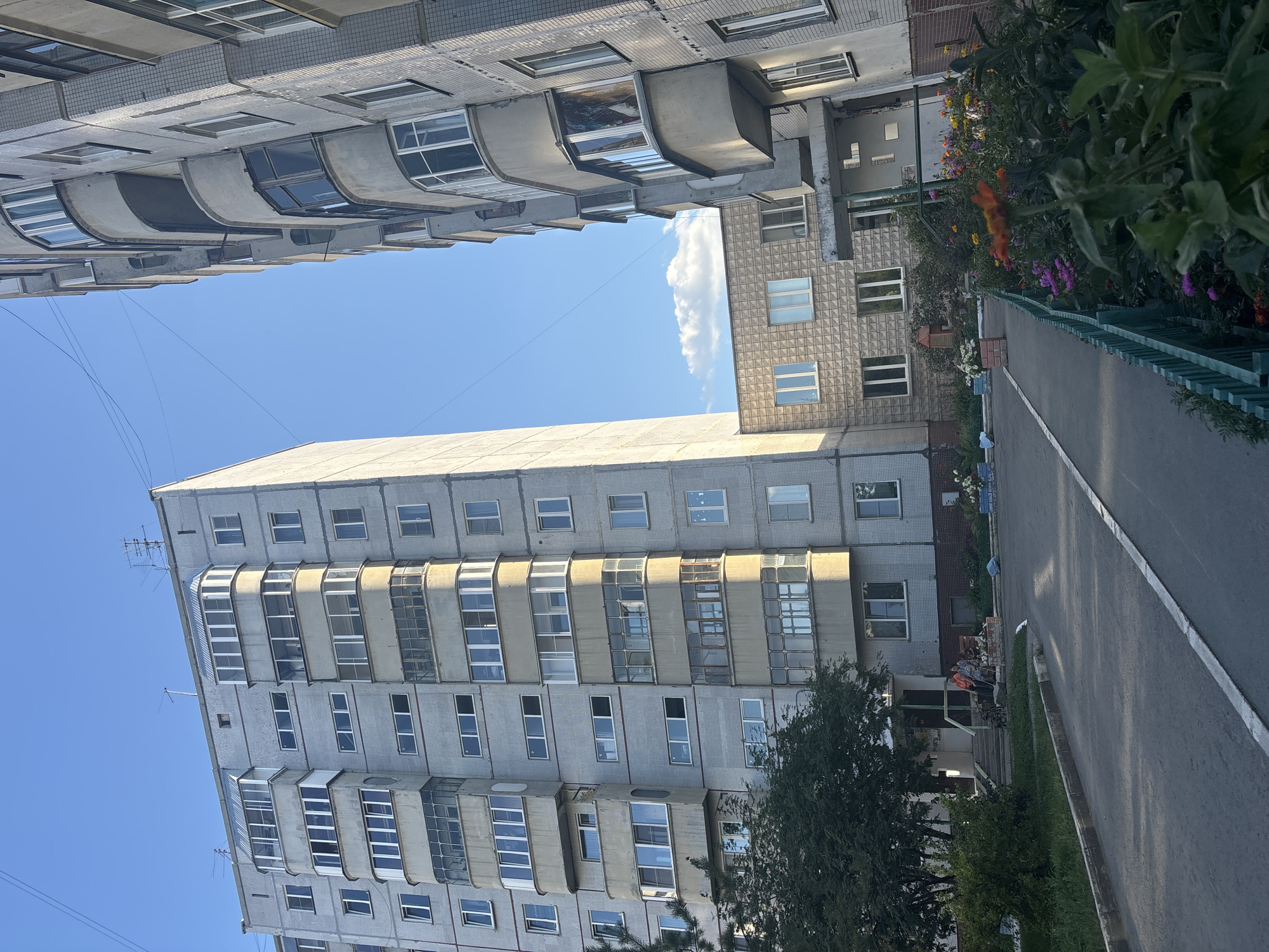 The image depicts a typical Soviet-era apartment complex in the city of Ob, Russia. The buildings are constructed from concrete and grey brick, with numerous balconies and windows. A paved walkway with a white line runs through the foreground, leading towards the entrance of one of the buildings. A lush green lawn and flowerbeds with colorful flowers line the walkway, bordered by a green fence. Two individuals are seated on a bench near the entrance of the building, one appears to be an elderly person. The sky is clear blue with a few scattered clouds. The overall impression is one of a residential area in a Russian city, characterized by functional architecture and well-maintained green spaces.