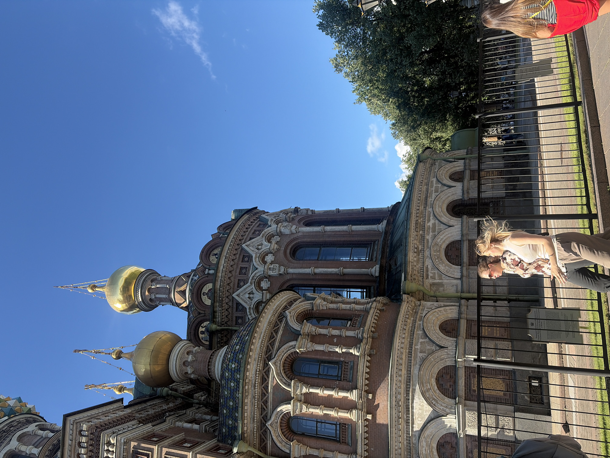 The image shows a side view of the Church of the Savior on Spilled Blood in Saint Petersburg, Russia, under a clear blue sky. The ornate architecture of the church, with its red brick facade, golden domes, and intricate detailing, is prominently featured on the left side of the frame. A black metal fence runs across the foreground, separating the sidewalk from the church grounds.
On the sidewalk, two people are walking from left to right. The person on the left is a woman wearing sunglasses, a patterned blouse, and dark pants. The person to her right is a woman with blonde hair wearing a sleeveless top and light-colored pants. Their postures suggest they are engaged in conversation. Further to the right, a woman with long brown hair is walking away from the camera, wearing a striped top and a red skirt or shorts.
Behind the fence, on the church grounds, there are glimpses of trees and more of the church's structure. The environment appears to be a public space, likely a tourist area, given the presence of visitors and the well-maintained appearance of the church and its surroundings. The lighting indicates it is daytime, with sunlight illuminating the scene.