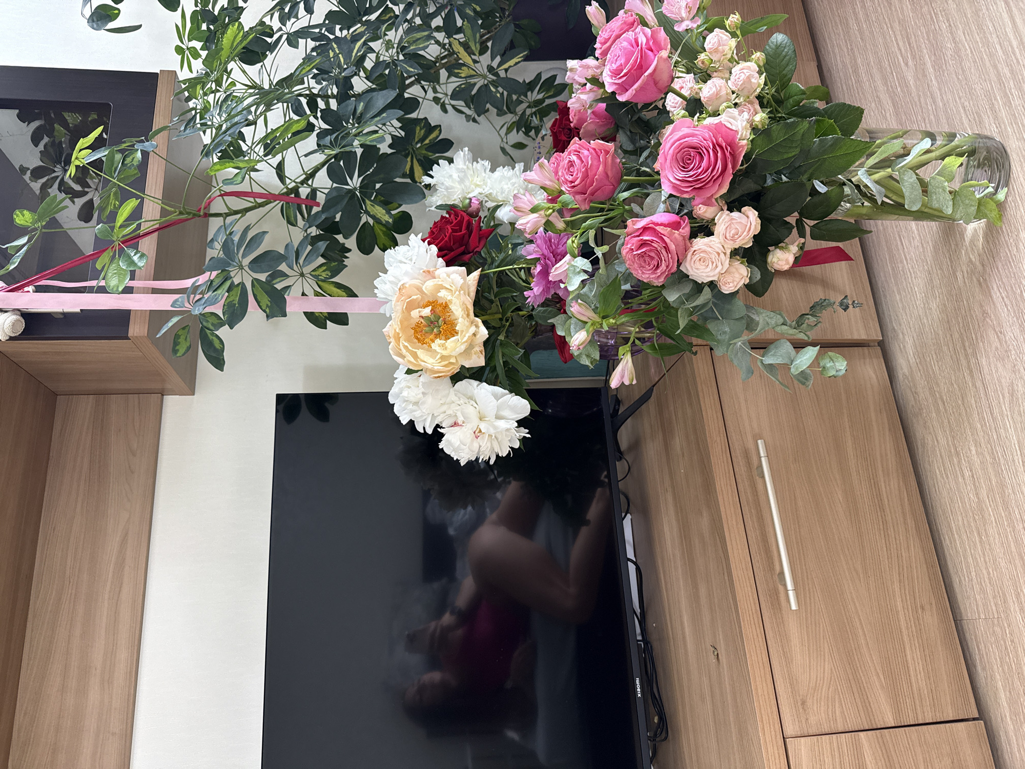 A high-angle, full shot captures a vibrant bouquet of flowers displayed on top of a television in an indoor setting. The bouquet is a colorful arrangement of pink roses, white peonies, red roses, and delicate pink alstroemeria, nestled amongst lush green foliage. Some of the flowers appear to be budding while others are in full bloom. A clear glass vase holds the bouquet, with stems visible inside. A thin, pink ribbon is tied around the foliage, and another pink ribbon is draped across the branches of a large plant in the background, possibly a Schefflera.
The television is a large, black, flat-screen model with a reflective surface that shows a distorted image of what appears to be a person wearing a dark pink top. The television is placed on a wooden entertainment unit. To the left of the television is a section of the same unit, with a darker frame and a glass-fronted cabinet. To the right of the television, more of the wooden entertainment unit is visible, featuring a cabinet with a brushed metal handle. The wall behind the bouquet is a plain white. The overall impression is one of domestic elegance, with the floral arrangement serving as a striking focal point. The lighting suggests it is daytime, and the setting appears to be a living room or similar common area within a home. There is no visible text in the image.