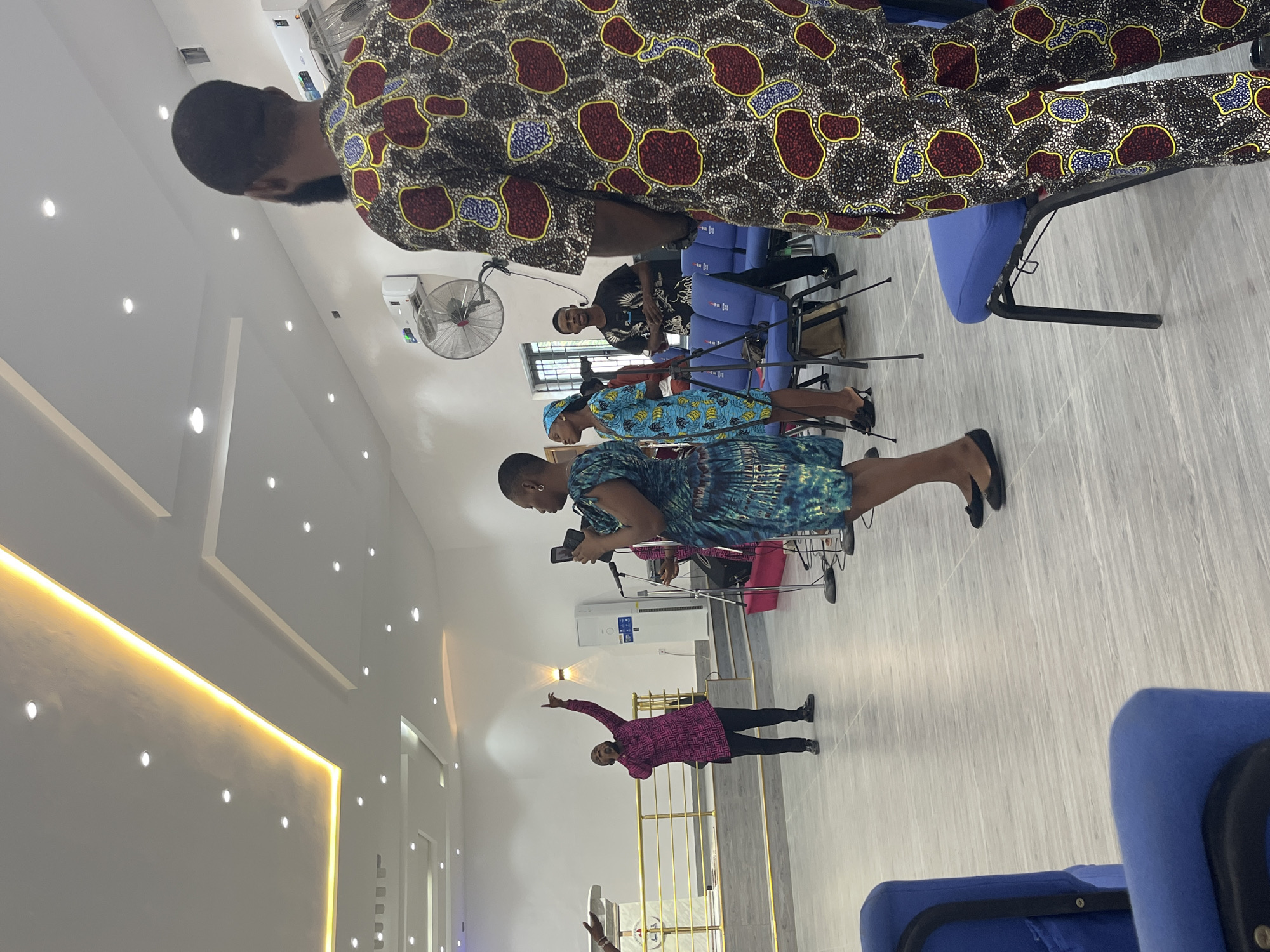 The image depicts an indoor scene, likely a church or conference hall in Ado, Nigeria. A woman in a blue and green dress is holding a smartphone, possibly recording or taking pictures. In front of her, another woman in a pink top and black pants stands with her arms raised. Several other people are present, seated on blue chairs, with a man standing behind the seated group. A man in a patterned brown top stands prominently in the foreground. The environment includes a white wall with a yellow light strip and a fan is visible on the wall.
