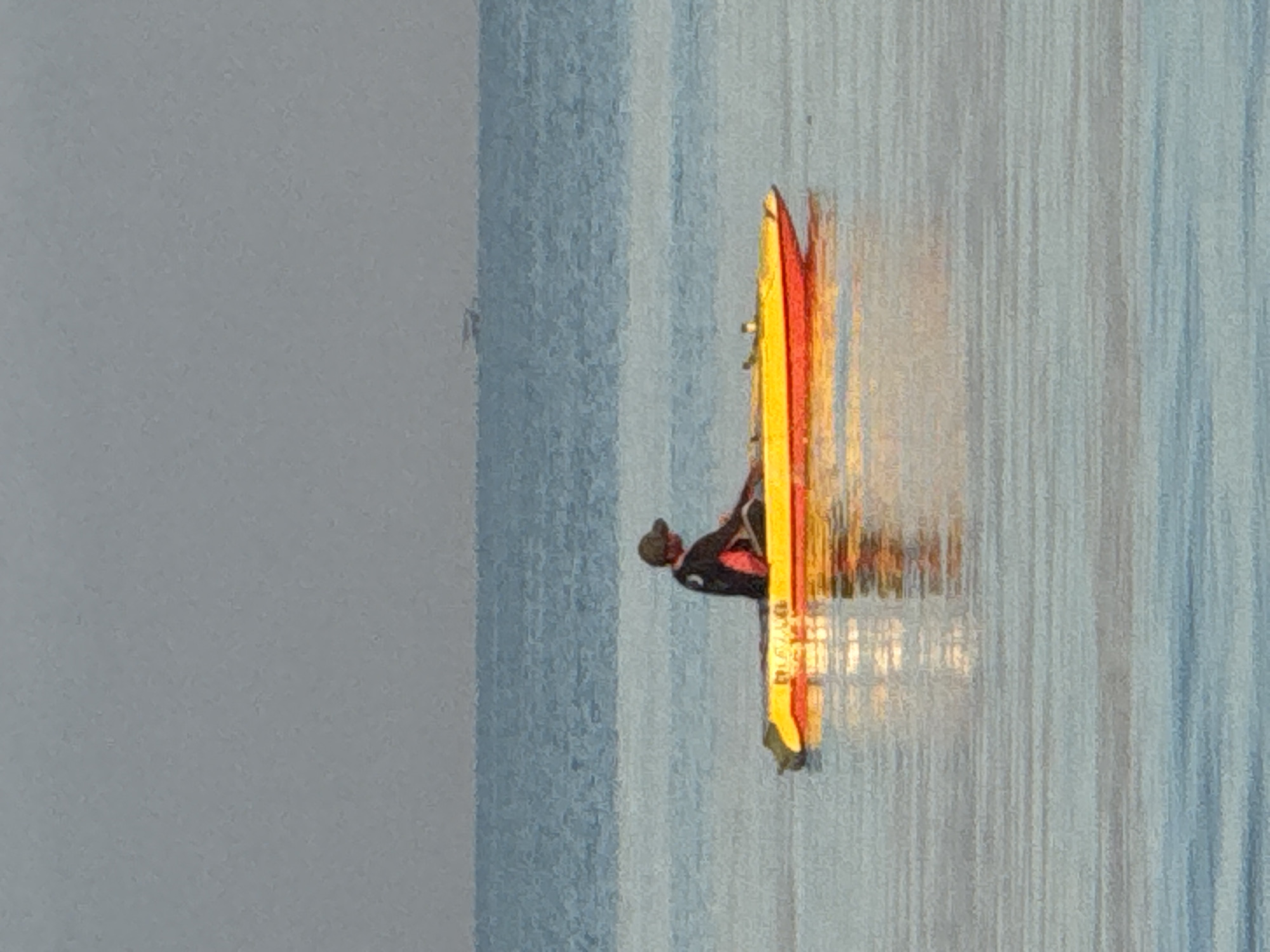 A person in dark clothing and a baseball cap stands on a yellow and red surfboard, close to a vertically oriented light blue wall that extends across the frame, suggesting an indoor setting in Jimbaran, Indonesia. The surfboard appears to reflect light, possibly from an interior light source. The background is a light gray.