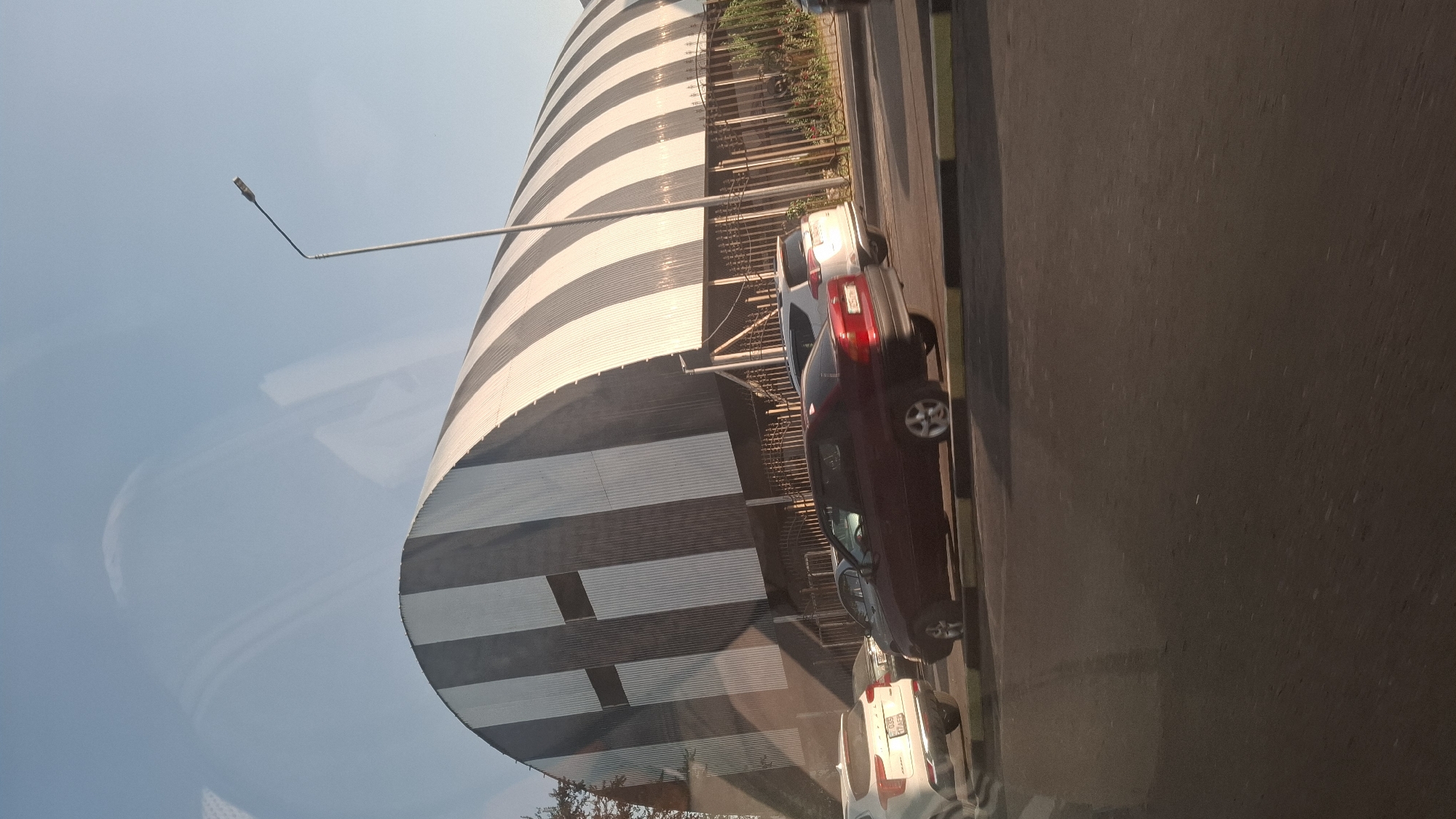 The image shows a street scene in Shymkent, Kazakhstan, with a distinctive arched building in the background. The building has a corrugated metal exterior with a pattern of alternating black and white vertical stripes, and a large dark rectangular shape on its front. In the foreground, several cars are visible on a road. A dark red sedan is in the middle of the frame, and a white SUV is to its left. Further back, another white car is parked. The street has a yellow and black striped curb. A tall street lamp stands on the right side of the frame. The sky is clear and blue, with some reflections visible on what appears to be a window. The overall scene suggests a daytime view of an urban environment.