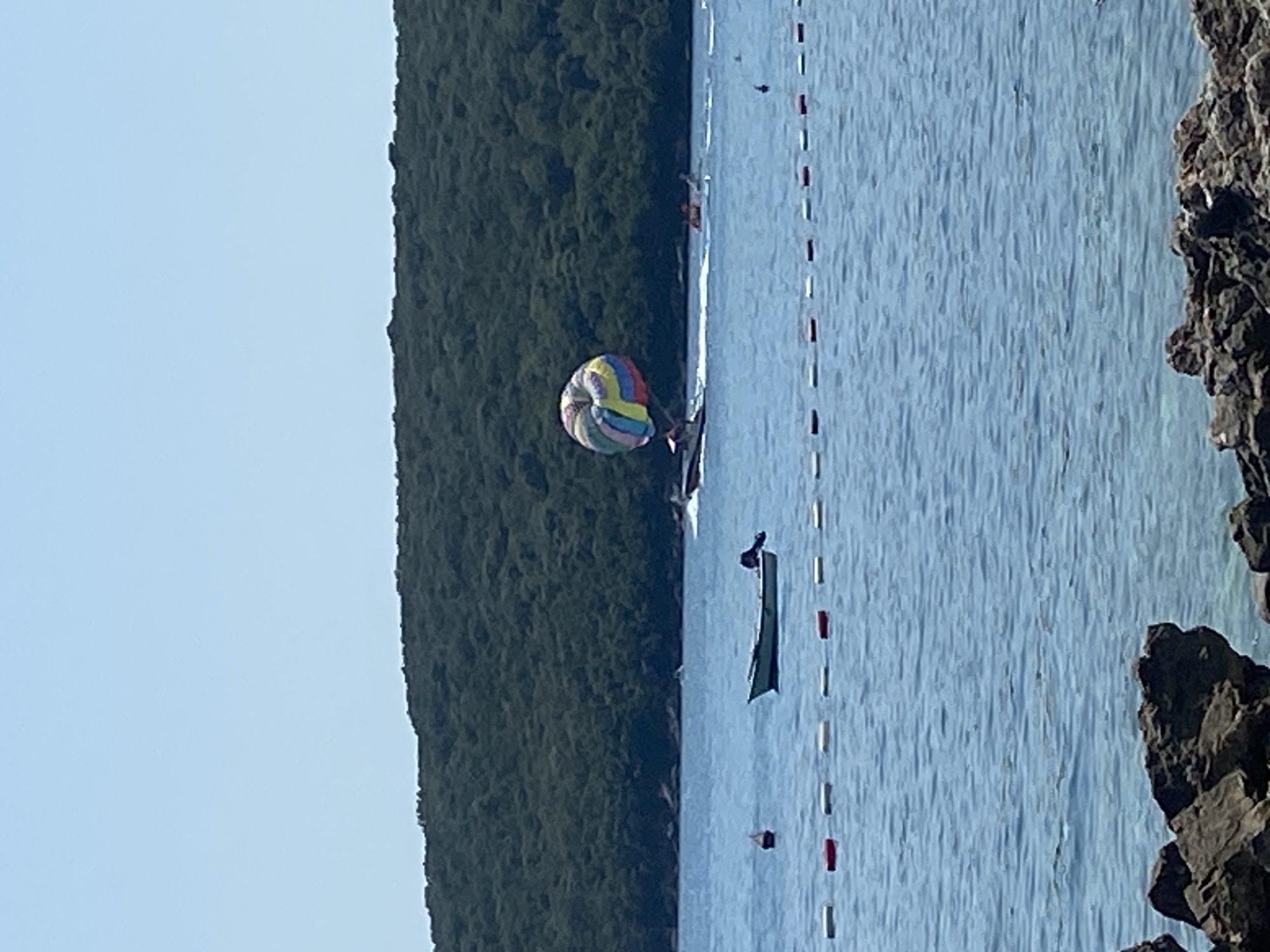 The image depicts a bright, sunny day at a coastal location in Prijevor, Montenegro. The scene is dominated by a calm, blue sea that stretches towards a heavily wooded, green hillside in the background. In the foreground, the edge of a rocky shore is visible. A line of buoys, alternating red and white, marks a boundary in the water.
In the middle ground, a small, light-colored boat with a motor is anchored. Further out, a parasailor is being pulled by a boat, with the colorful parachute high in the sky. Another person appears to be in the water near the parasailing boat, possibly waiting their turn or having fallen. Additional boats are visible in the distance, some creating small wakes on the water. The overall atmosphere suggests a popular recreational area, likely a beach or a bay.