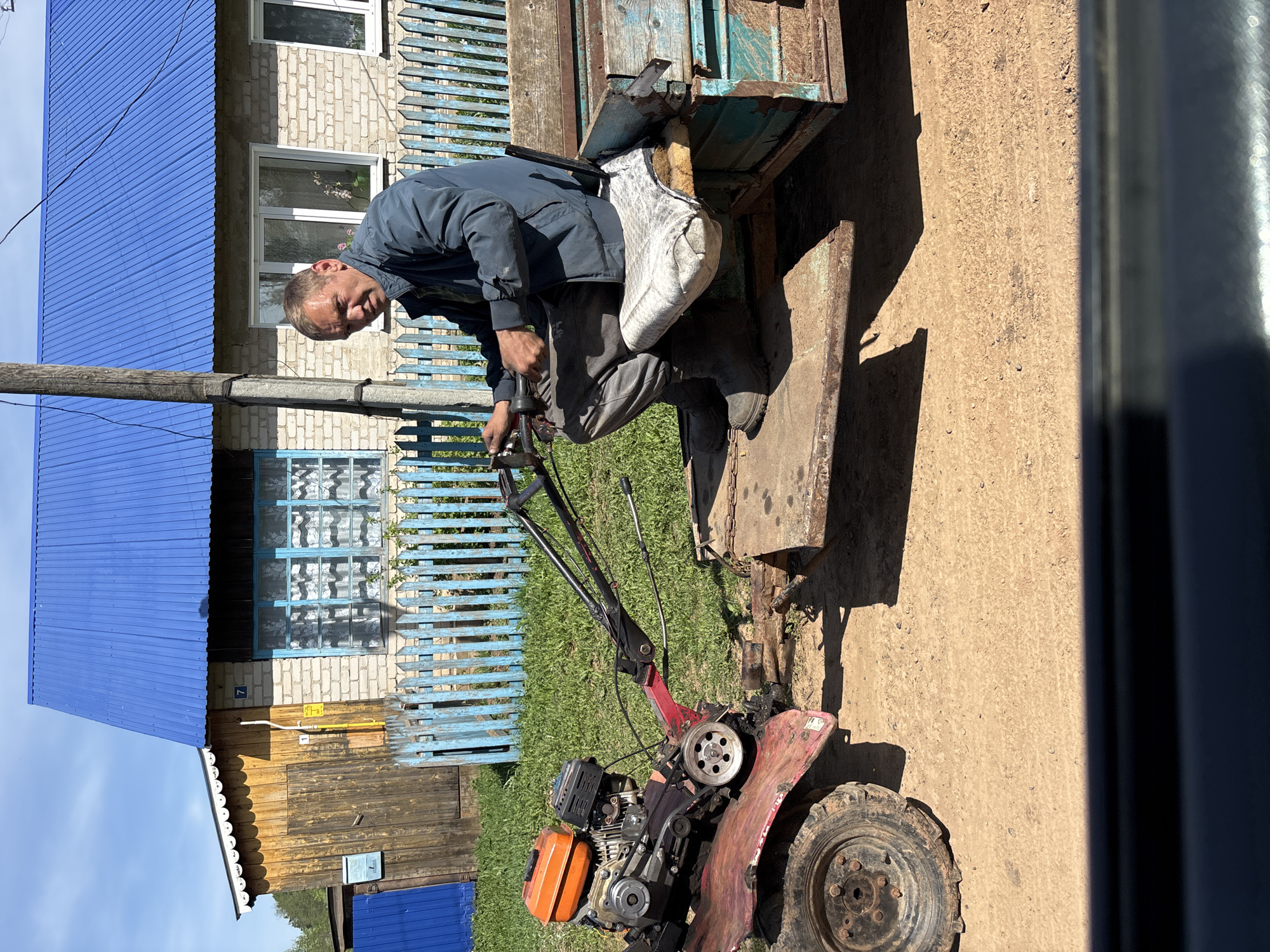 The image depicts a man in Markidonovka, Russia, working on a small tiller-like agricultural machine attached to the back of a truck or trailer. He is wearing a dark jacket and grey pants, and is crouched, appearing to manipulate controls of the machine. The machine is red and black, with an orange fuel tank. It stands on dirt ground near a light-blue picket fence and building with a blue metal roof.