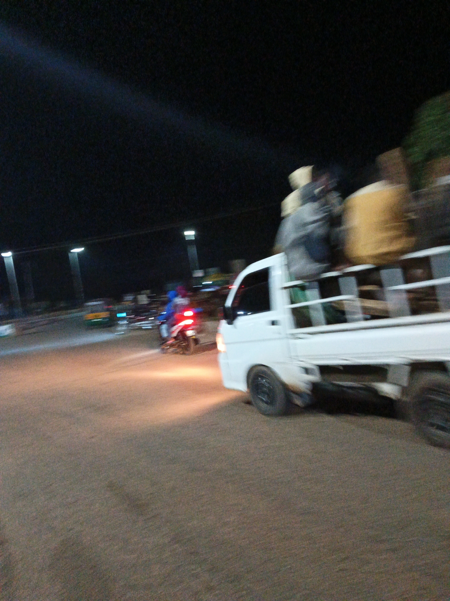 In Bukuru, Nigeria, a white pickup truck is captured in motion at night. The truck is loaded with several individuals and what appear to be large, cylindrical objects wrapped in yellow material. The individuals are blurry due to the motion, but at least three are discernible. The truck is traveling on a paved road, illuminated by streetlights and the headlights of other vehicles. In the background, a motorcycle with a rider is also in motion, its red taillight visible. Other vehicles and streetlights can be seen in the distance. The overall scene depicts a busy nighttime street in Bukuru.