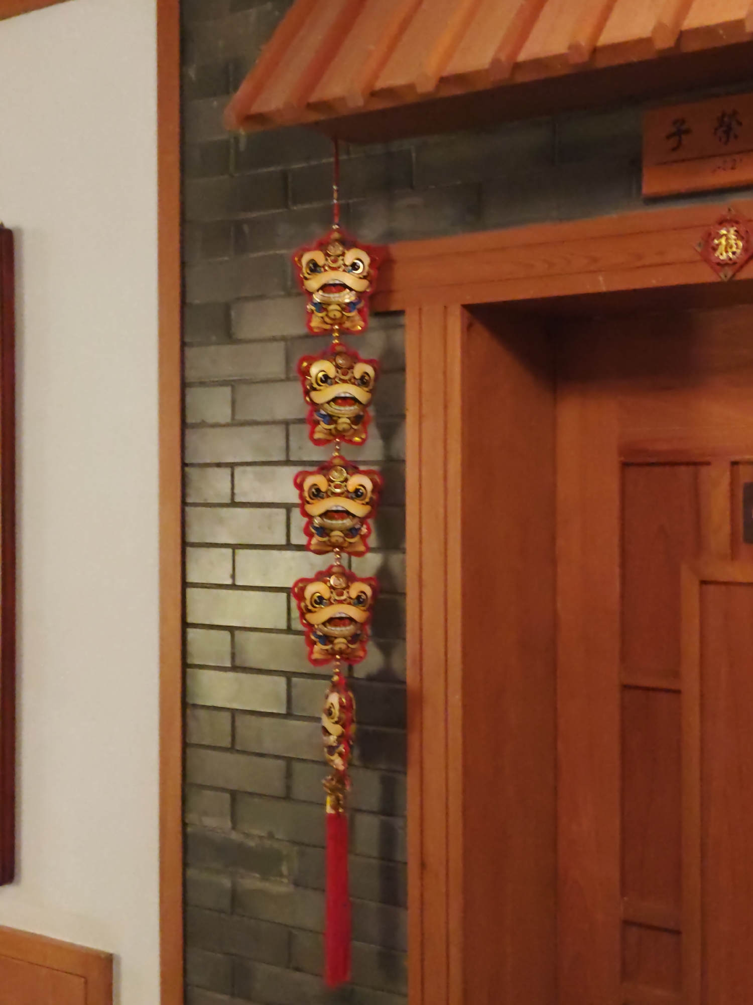 A string of five red and gold lion head ornaments hangs vertically against a grey brick wall. The ornaments are spaced evenly, with the bottom one featuring a red tassel. To the right, a wooden door with vertical panels is visible. Above the ornaments and door, a wooden eave with orange slats is present, and a small wooden plaque with Chinese characters is mounted on the brick wall. The left side of the image shows a white wall with a wooden trim. The setting appears to be indoors, possibly a building entrance or hallway. The location is Lindian, China.
