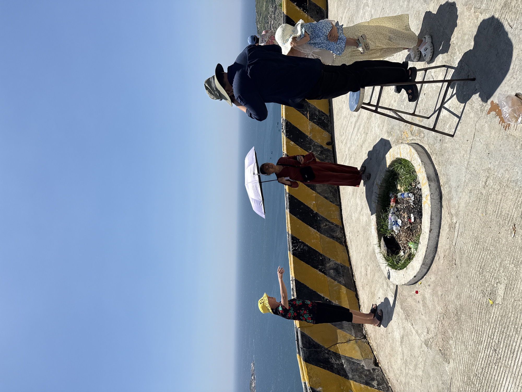 The image captures an outdoor scene at a coastal overlook in Vinh Hai, Vietnam, under a clear, bright blue sky. A group of five adults and one child are on a concrete platform, which is bordered by a concrete barrier painted with black and yellow diagonal stripes.
On the left, an adult, likely female, wearing a yellow bucket hat, a dark floral shirt, and dark pants, points with her right arm towards the sea. To her right, another adult, likely female, in a long red dress, holds a white umbrella overhead for shade and carries a dark shoulder bag. Further to the right, an adult, likely male, in a dark green wide-brimmed hat and a dark blue long-sleeved shirt, stands elevated on a light blue metal stool, with his back partially to the viewer, appearing to view or operate an object held to his face. Adjacent to him, an adult, likely female, in a light-colored long skirt and top, holds a small child dressed in a blue patterned outfit. Another adult in a light shirt and blue hat is partially visible in the background behind the man on the stool, also facing the sea.
In the foreground, a circular concrete-lined pit contains sparse green vegetation and accumulated refuse, including plastic bottles and other debris. Near the base of the stool, a clear plastic bag containing a brown substance appears to have spilled onto the concrete surface. The background features a wide, clear blue sky meeting a calm, deep blue sea, with small rocky islands visible in the distance on the left and several faint boat silhouettes on the water.