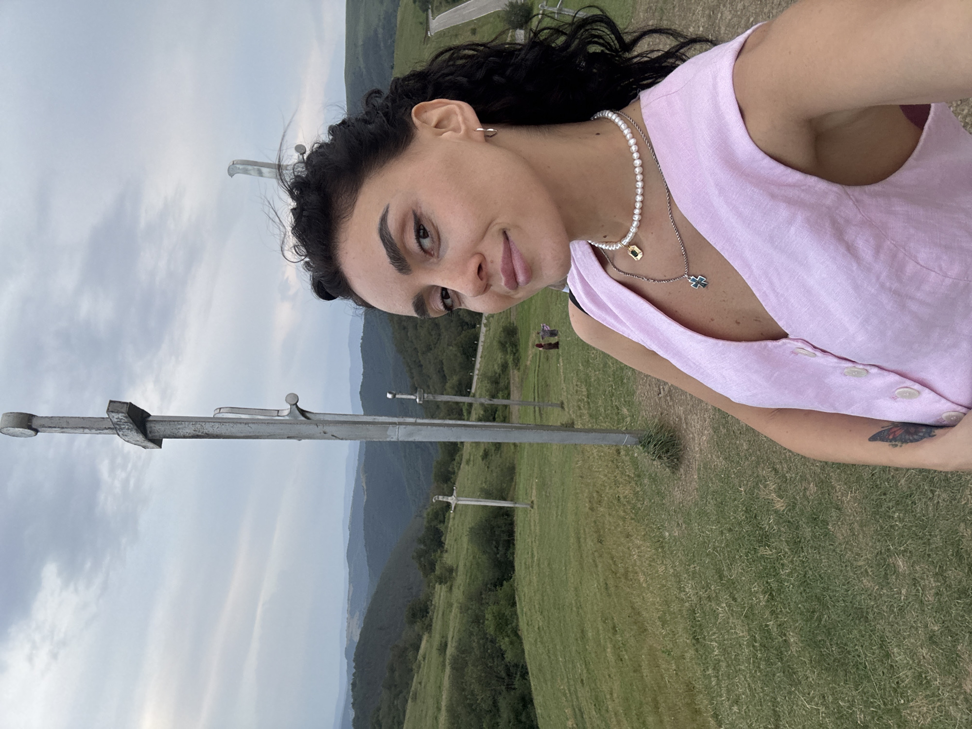 A selfie is taken by a young woman with dark, curly hair, wearing a light pink sleeveless top and a pearl necklace. She is standing outdoors in a hilly, grassy landscape with a cloudy sky. In the background, there are several large metallic sword-like structures planted in the ground, and a winding road is visible in the distance. The woman is smiling and looking towards the camera. The location is identified as the site of the "Swords of God" monument in Zemo Nichbisi, Georgia.