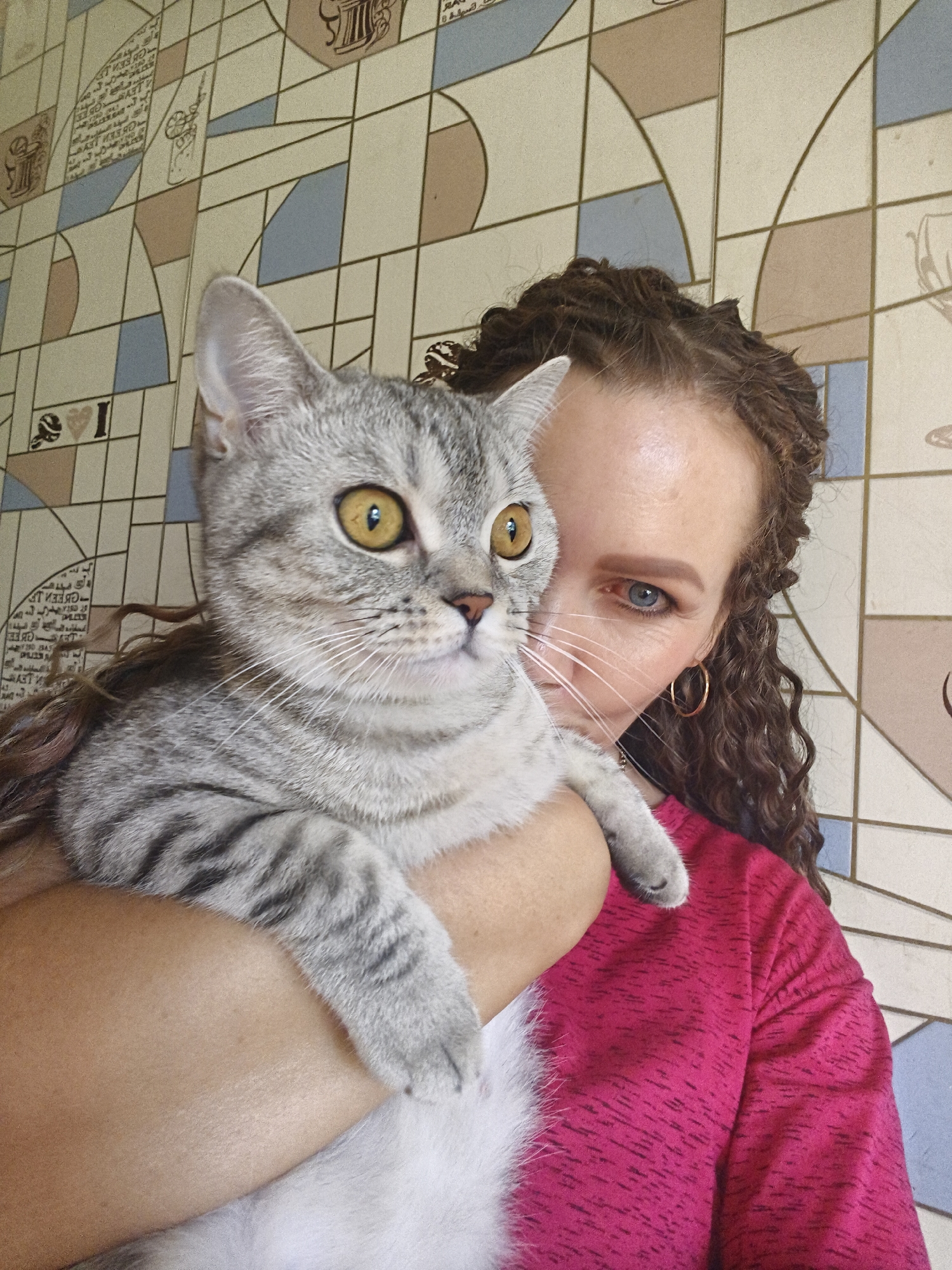 A woman with curly brown hair and blue-gray eyes holds a gray and white tabby cat in front of a tiled wall. The woman is wearing a pink shirt. The cat has large yellow eyes and is looking to the right. The wall has a geometric pattern of beige, blue, and pink tiles.