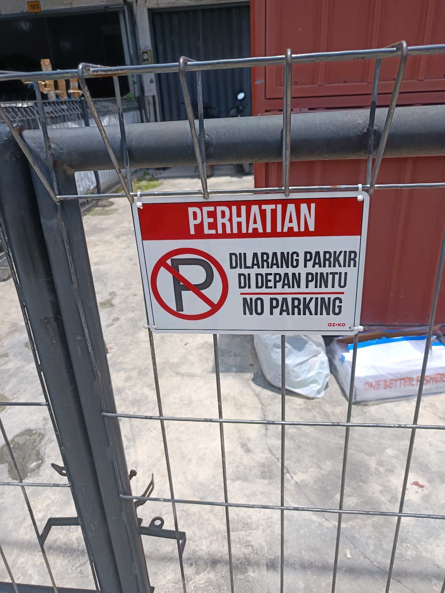 The image shows a red and white sign with "PERHATIAN" at the top, meaning "ATTENTION" in Indonesian. Below this, the sign reads "DILARANG PARKIR DI DEPAN PINTU NO PARKING," which translates to "DO NOT PARK IN FRONT OF THE DOOR NO PARKING." The sign is attached to a metal fence. Behind the fence, there is a red shipping container and a building with the number "103" visible. There are also two white plastic bags and a white box with blue trim on the ground. The area appears to be an outdoor space, possibly a street or alleyway, in Percut Sei Tuan, Indonesia. The ground is paved with concrete. The image is taken from a slightly low angle, looking up at the sign and fence.