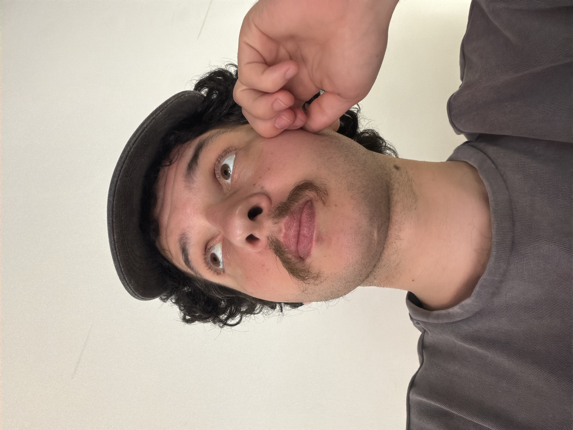 The image shows a close-up, tilted portrait of a young person with curly dark hair, wearing a dark cap and a grey t-shirt. Their face is turned towards the camera, with one hand reaching up to touch their cheek near the eye. The person has a surprised expression, with wide eyes and an open mouth. A thin mustache is visible above their upper lip. The background is a plain, light-colored wall, suggesting an indoor setting. The lighting is even, with no strong shadows, and there are no external environmental cues to indicate the time of day or weather. The image appears to be a selfie, captured from a low angle. There is no visible text in the image. The overall mood is playful and a bit startled.