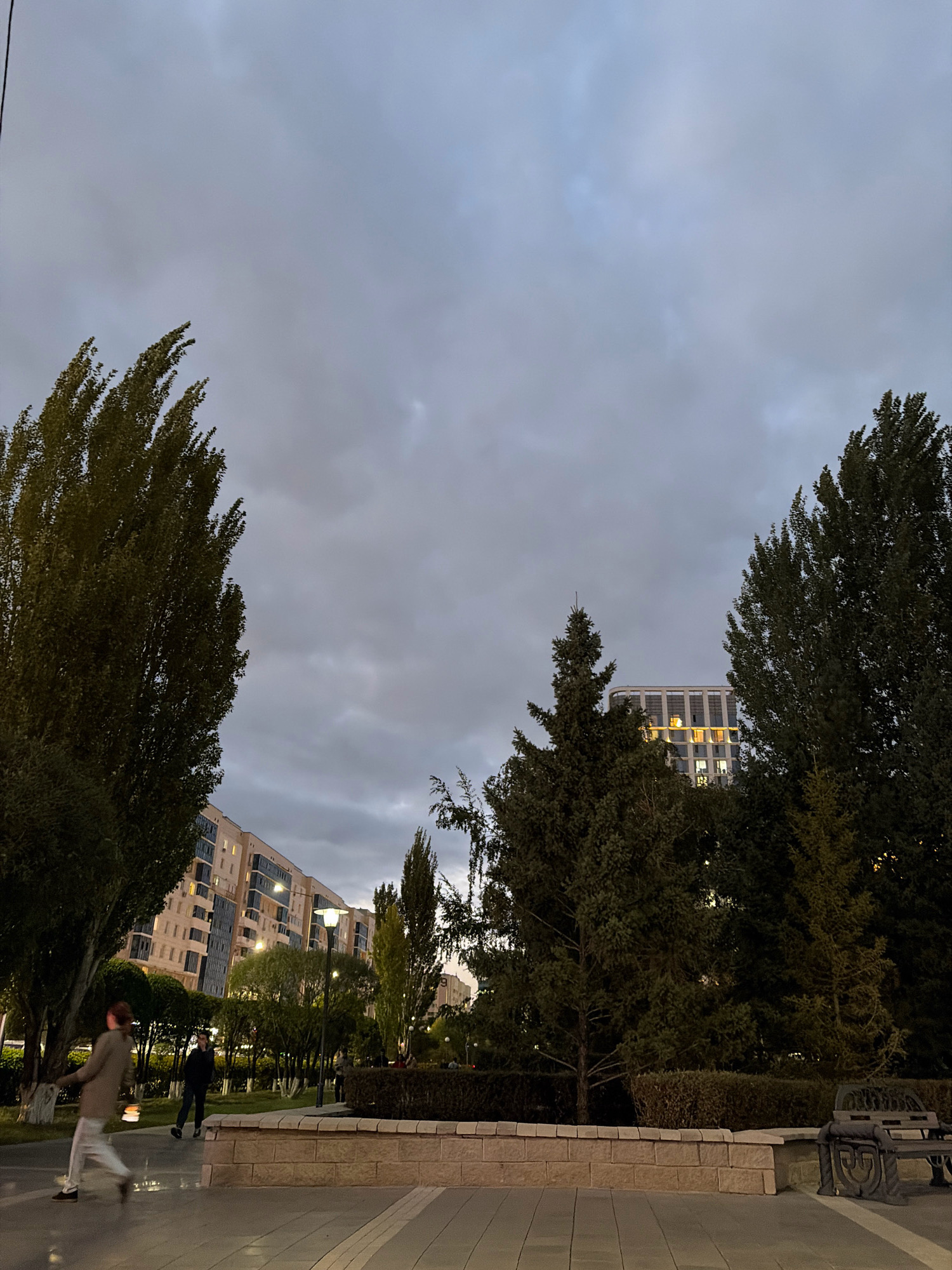 The image depicts a cloudy evening scene in a park-like area in Astana, Kazakhstan, possibly in a residential district. In the foreground, a paved walkway is visible with a low, brick-faced wall. To the right, a dark metal park bench sits on the walkway. Further back, a manicured area with bushes and trees is illuminated by lampposts. In the middle ground, two individuals are present: one person in the left foreground is walking with their arms outstretched, and another person stands further back in the middle of the park. A multi-story residential building, characterized by its light-colored facade and numerous windows with lights on, forms the backdrop to the park. Several tall, dark green trees are interspersed throughout the scene, framing the view of the building and contributing to the twilight atmosphere. The sky is overcast with thick, grey clouds.